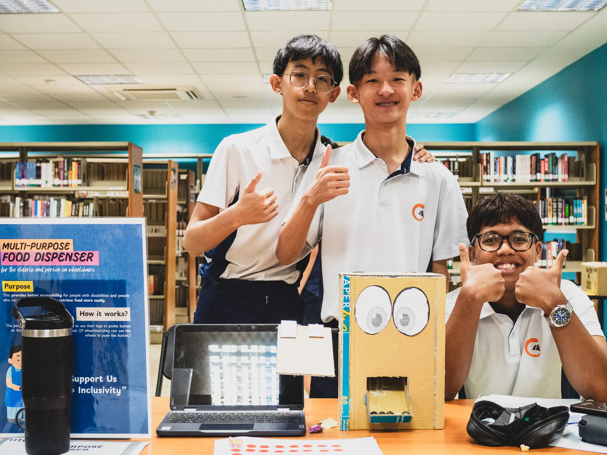 Three students give thumbs up beside a cardboard food dispenser model and a project poster in a school library.
