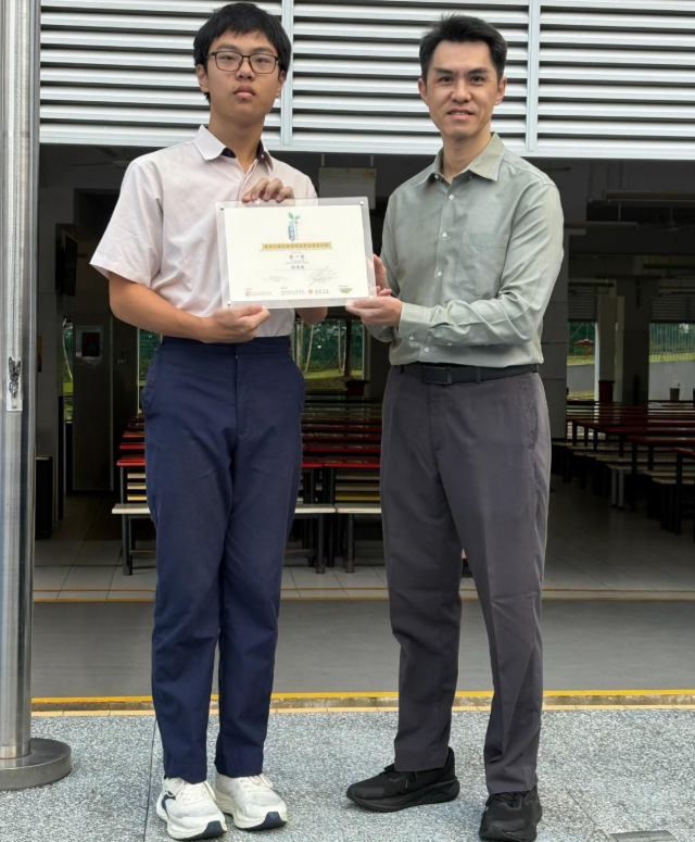 Two men stand together holding a certificate outdoors near a building with benches in the background.