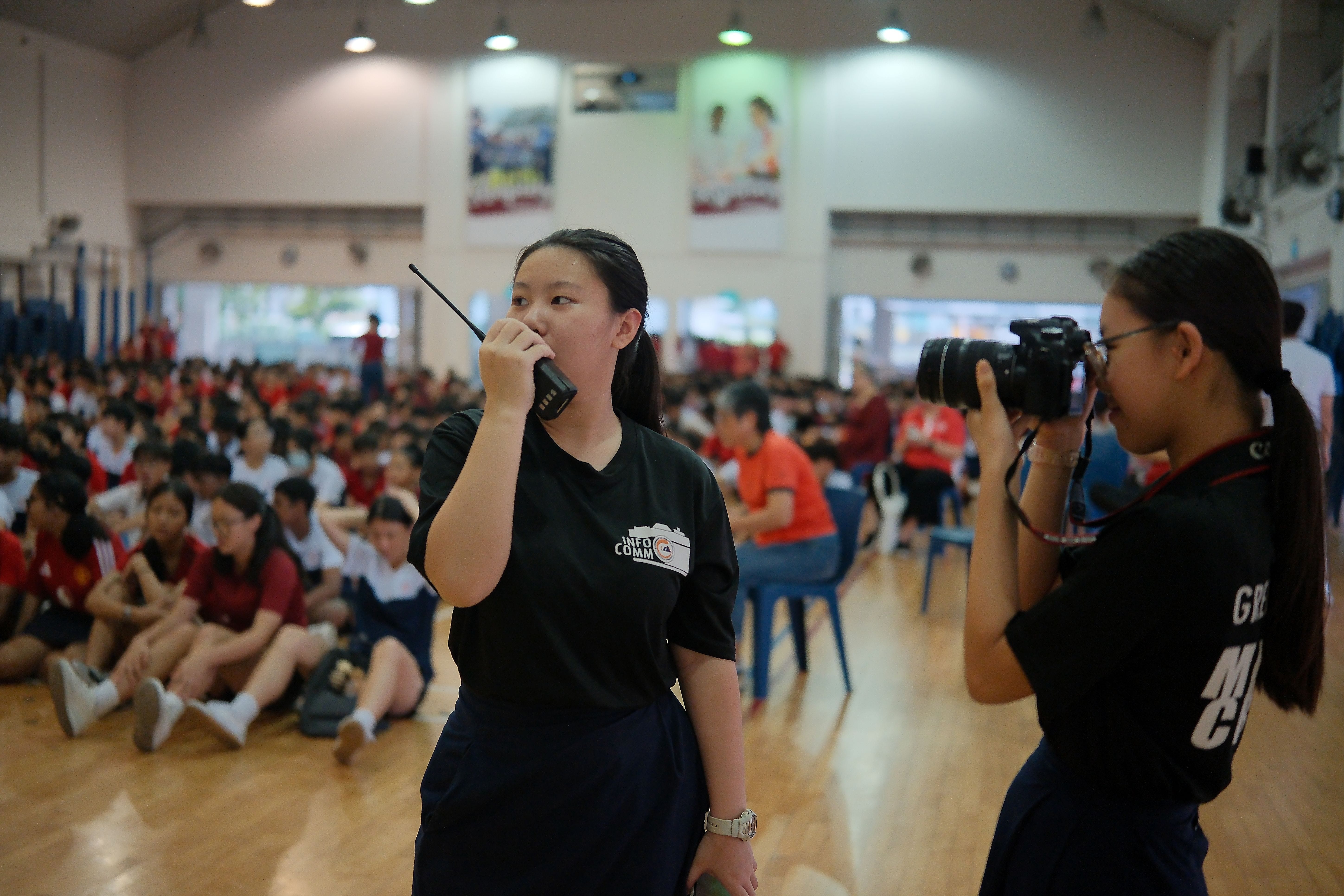 Two girls in black shirts work a school event; one speaks into a walkie-talkie, the other photographs with a DSLR camera.