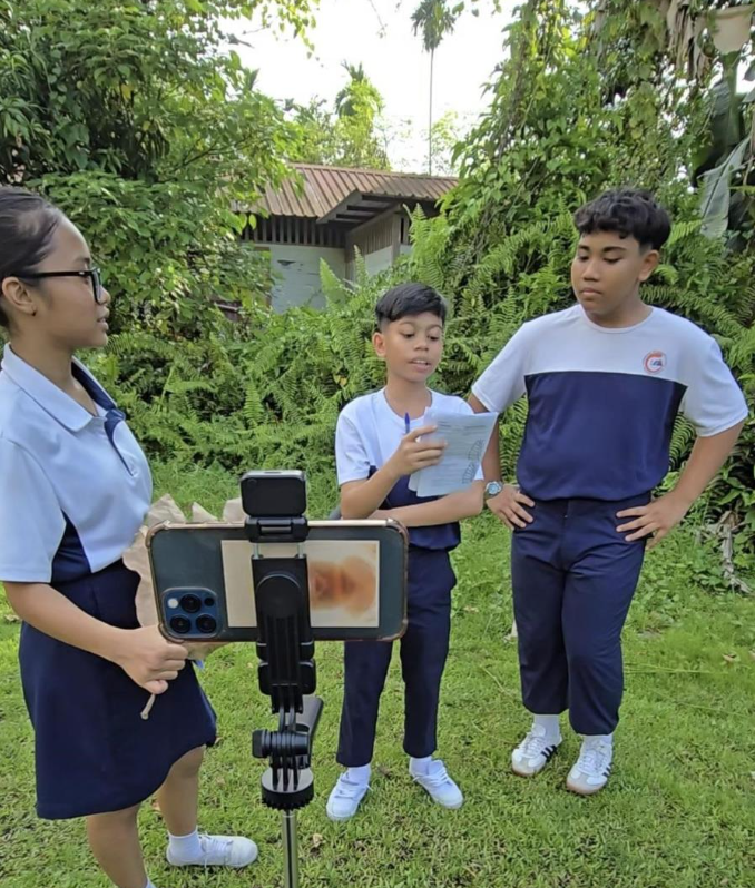 Three school students in uniform filming outdoors, one holding papers, with a phone on a tripod.