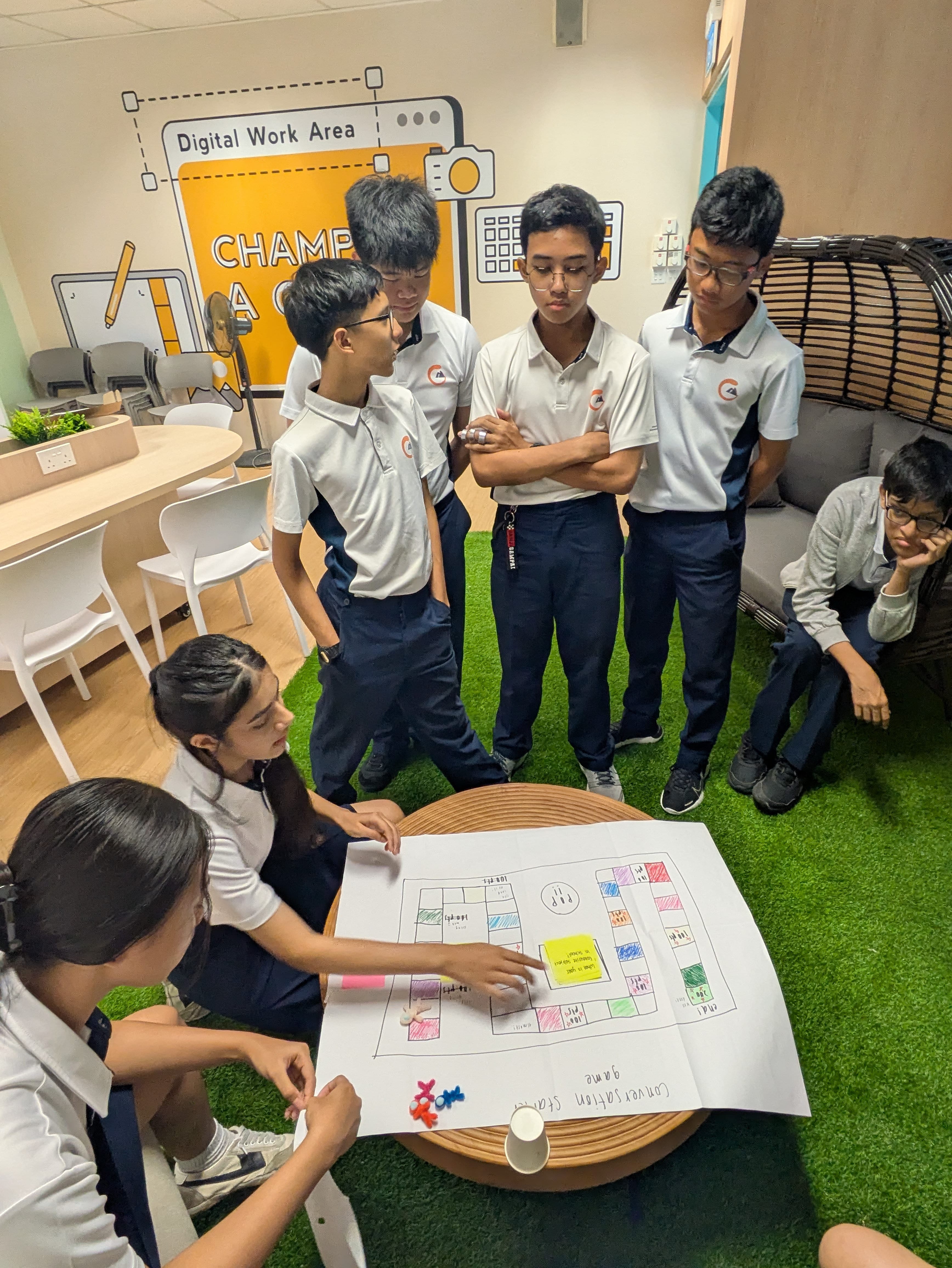 Students in school uniforms gather around a hand-drawn board game on a table in a Digital Work Area classroom.