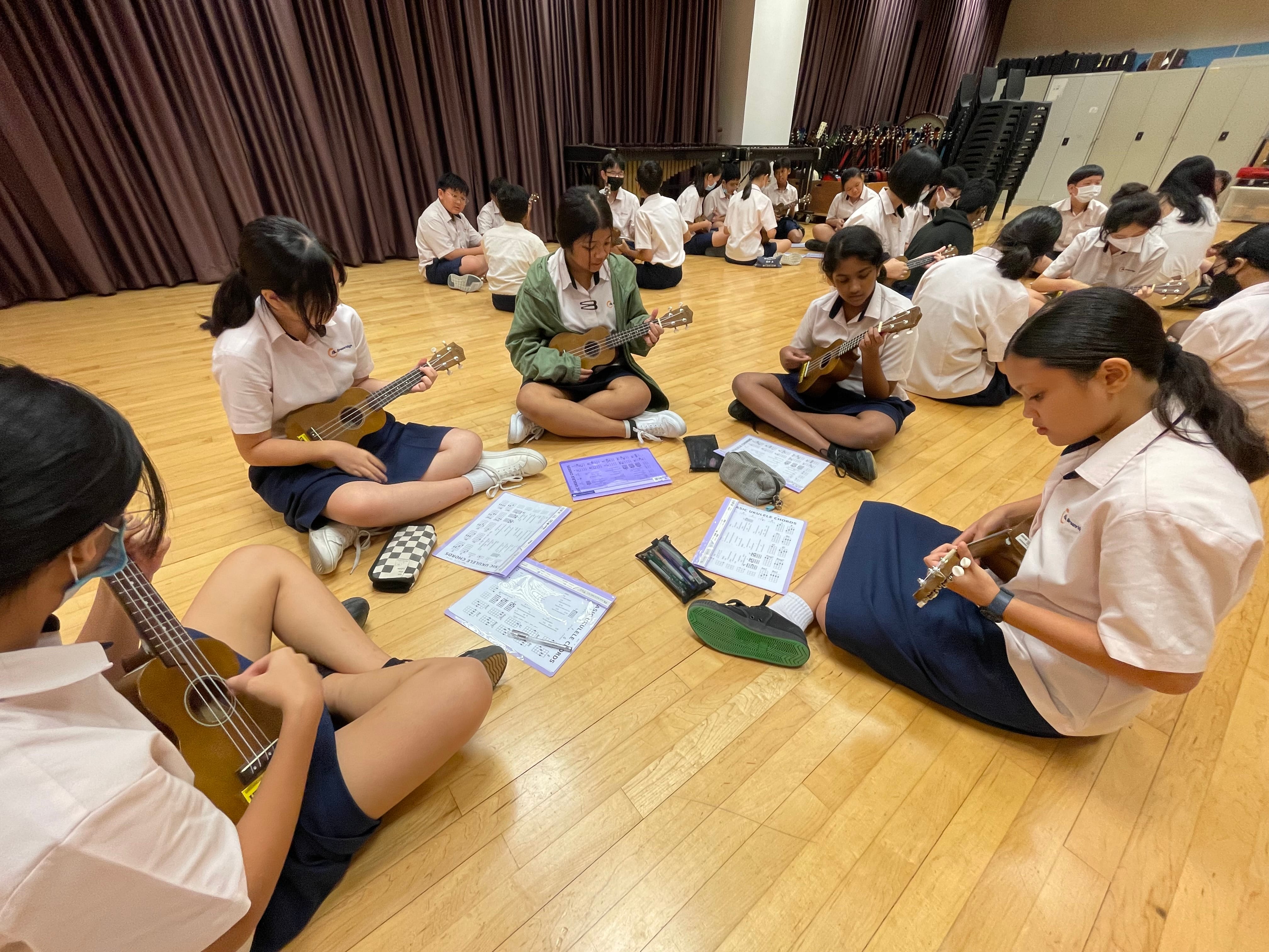 Students sit on a wooden floor in groups, practicing ukuleles with sheet music spread out.