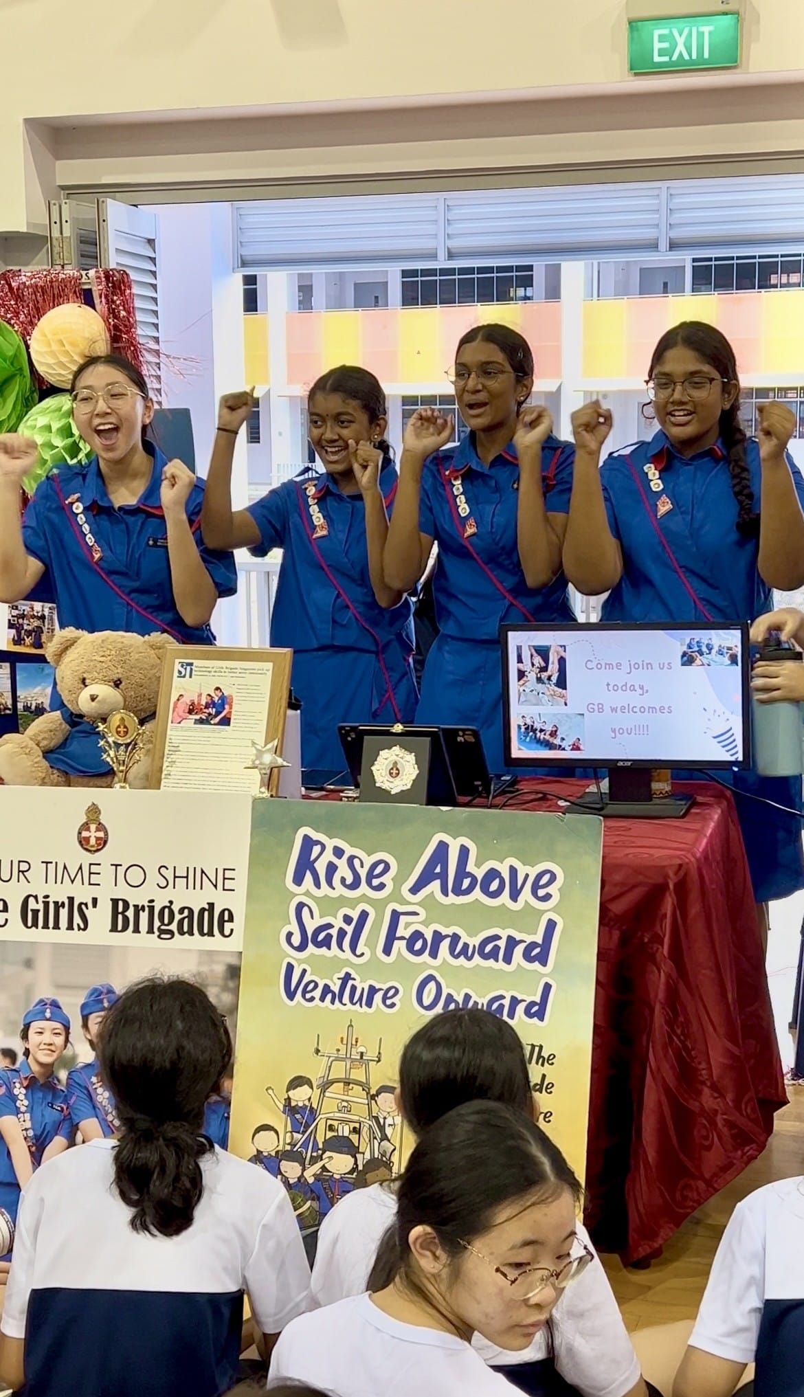 Four girls in blue Girls' Brigade uniforms cheer at a recruitment booth with signs reading Rise Above Sail Forward Venture Onward.
