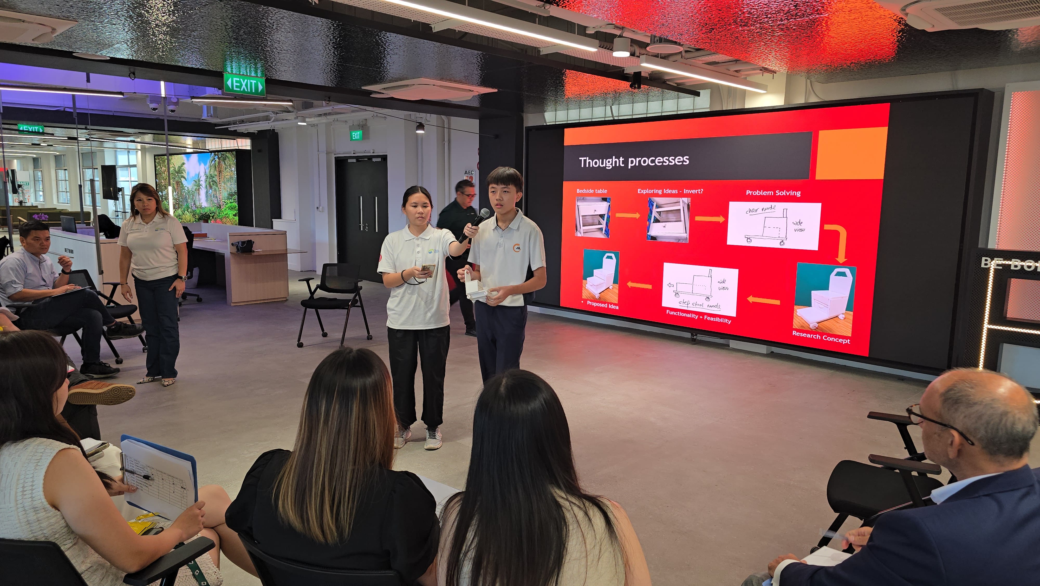 Two students present a "Thought processes" slide on a large red screen to a seated audience in a modern room.