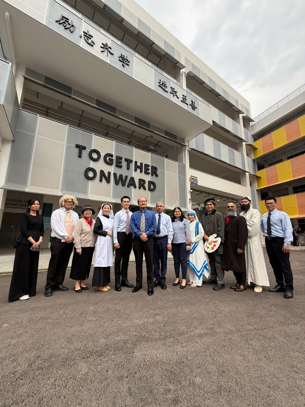 Group of people in various costumes standing in front of a building with "Together Onward" and Chinese text on it.