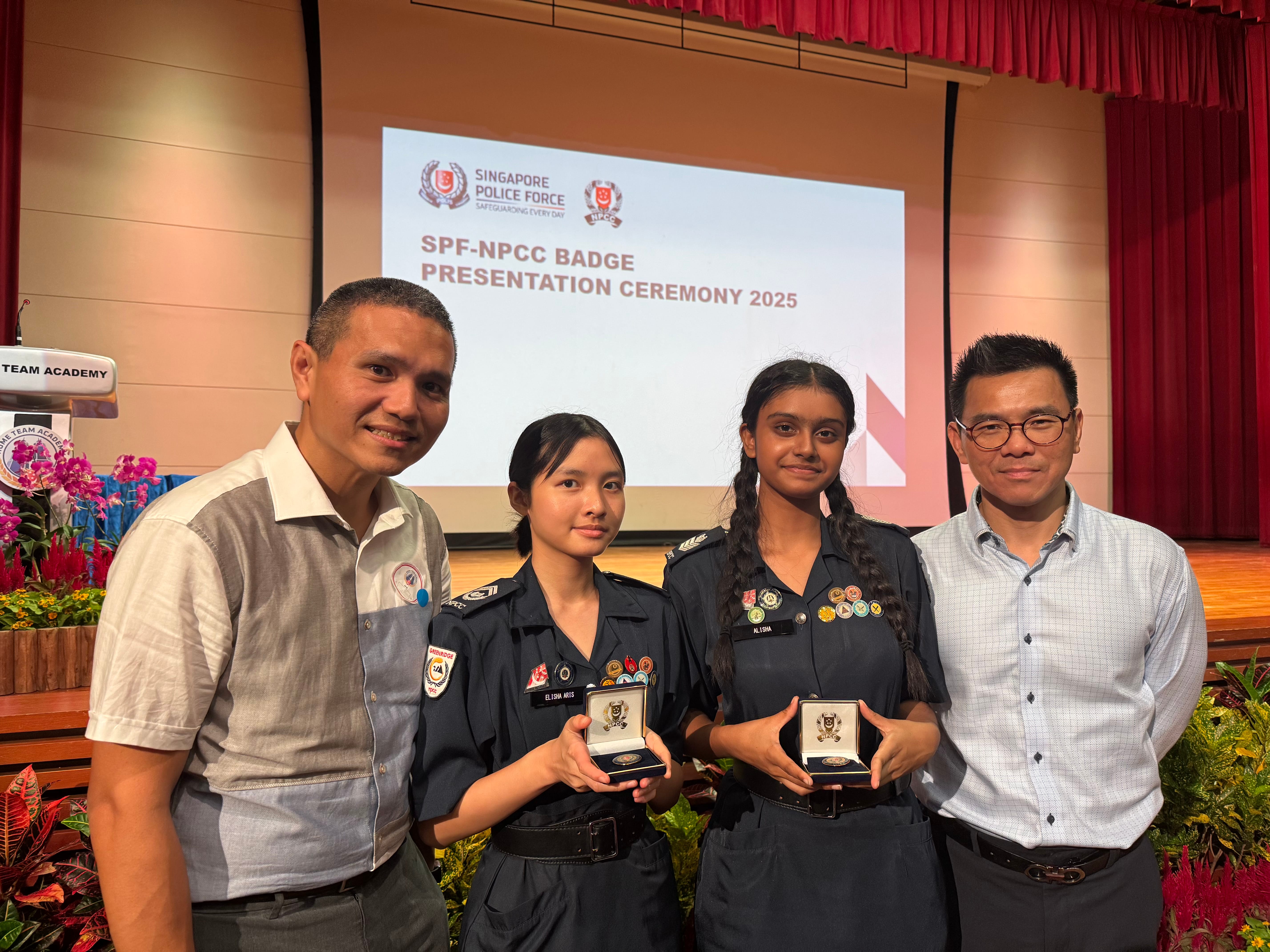 Four people pose onstage; two in uniform hold badge cases; screen reads SPF-NPCC Badge Presentation Ceremony 2025.