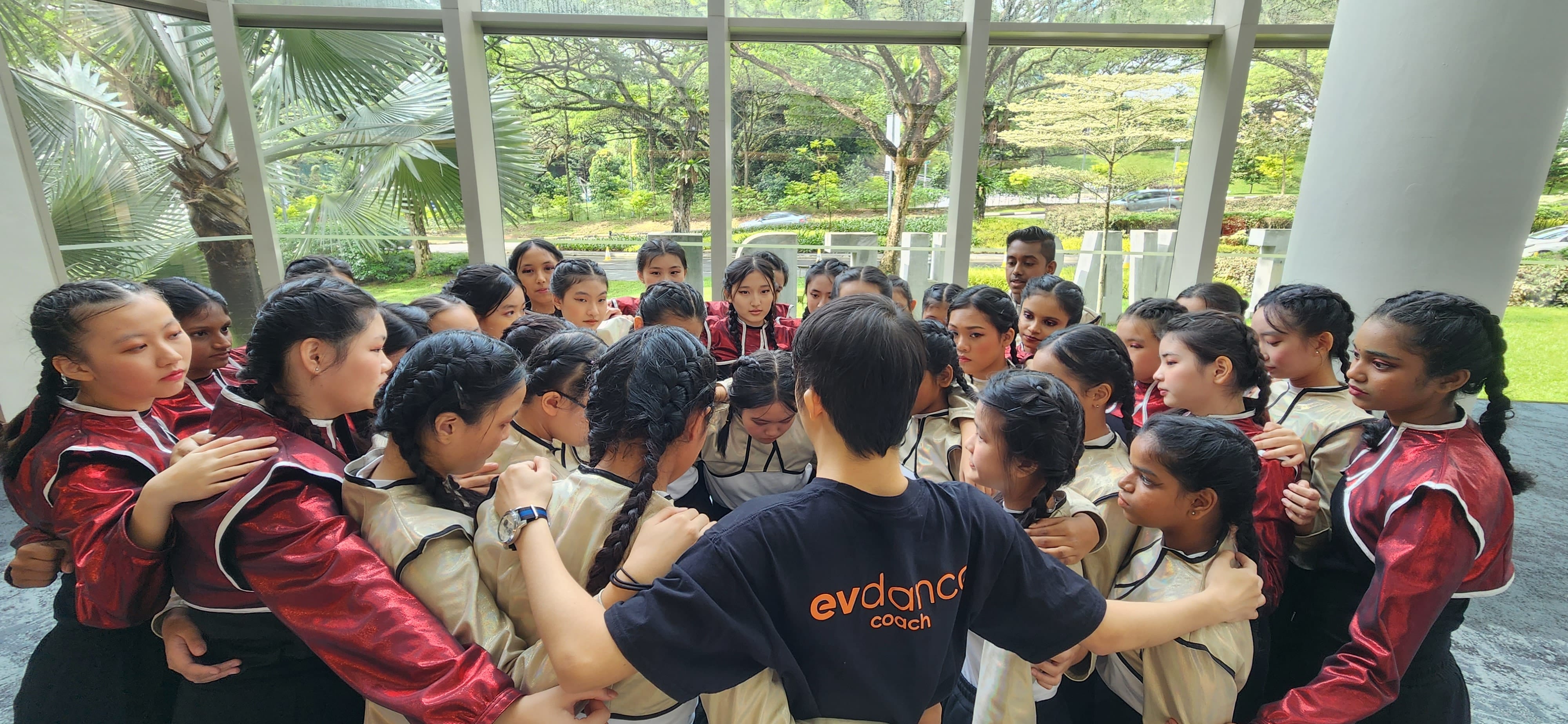 A dance coach in a navy evdance shirt huddles with a large group of young dancers in uniform indoors.