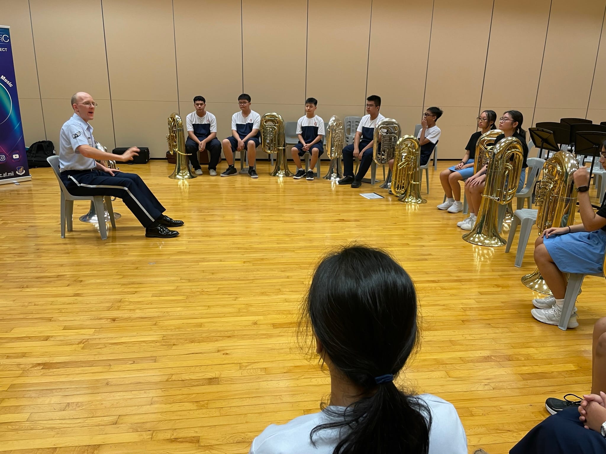 A uniformed instructor sits facing a semicircle of students with tubas on a wooden floor in a rehearsal room.