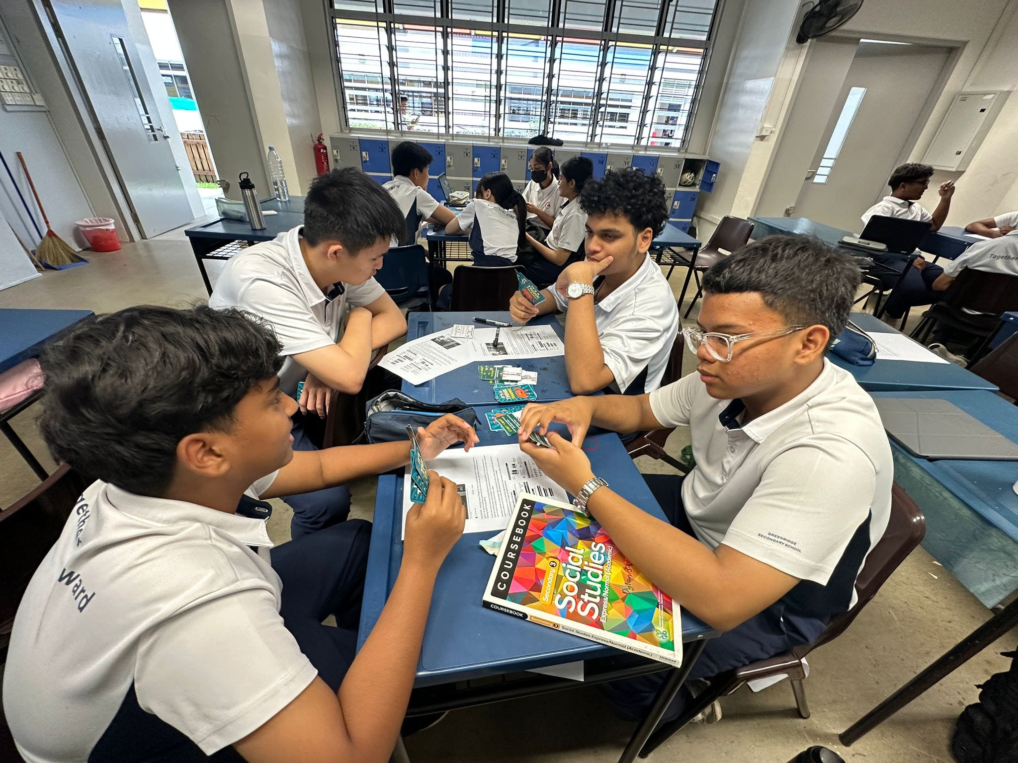 Three male students in white uniforms discuss around a desk with a Social Studies coursebook in a classroom.