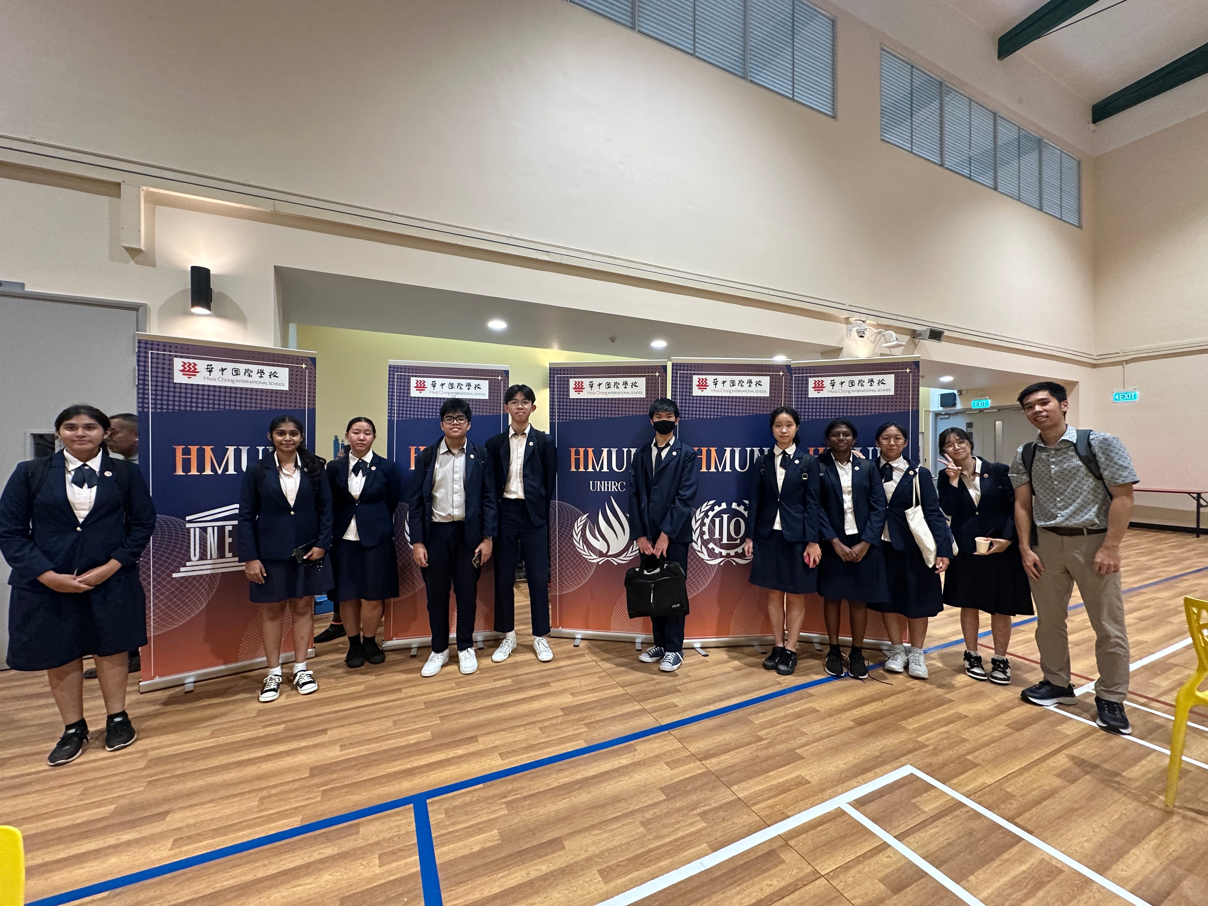 A group of students in navy uniforms stand before HMUN event banners inside a school hall with a wooden floor.