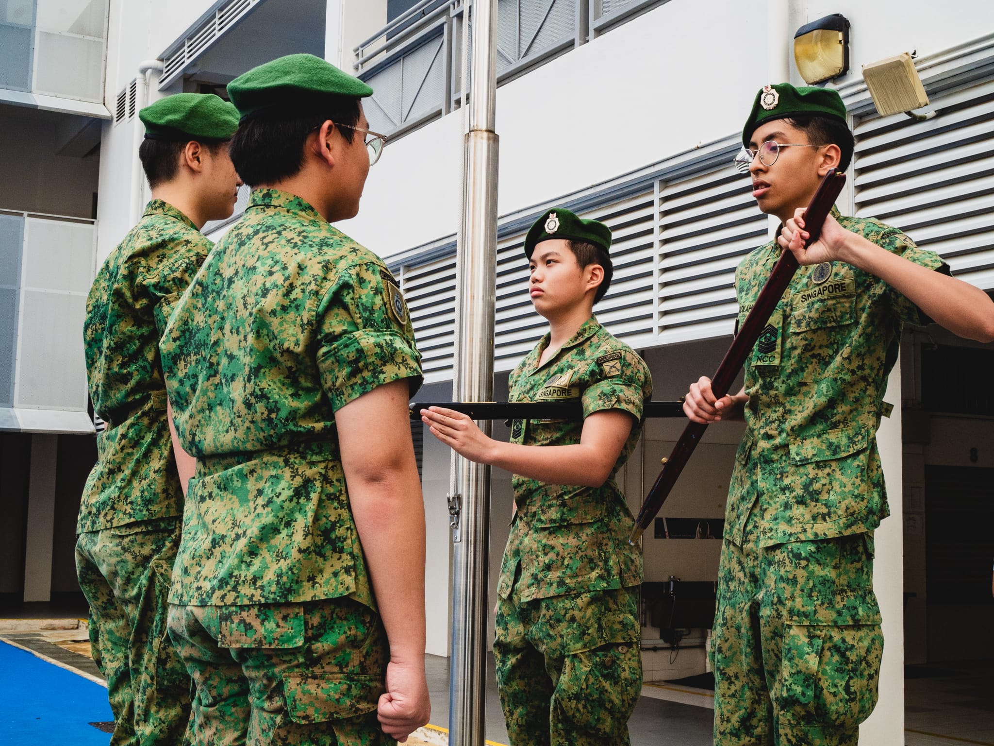 Four young people in Singapore NCC camouflage uniforms and green berets standing near a flagpole outdoors.