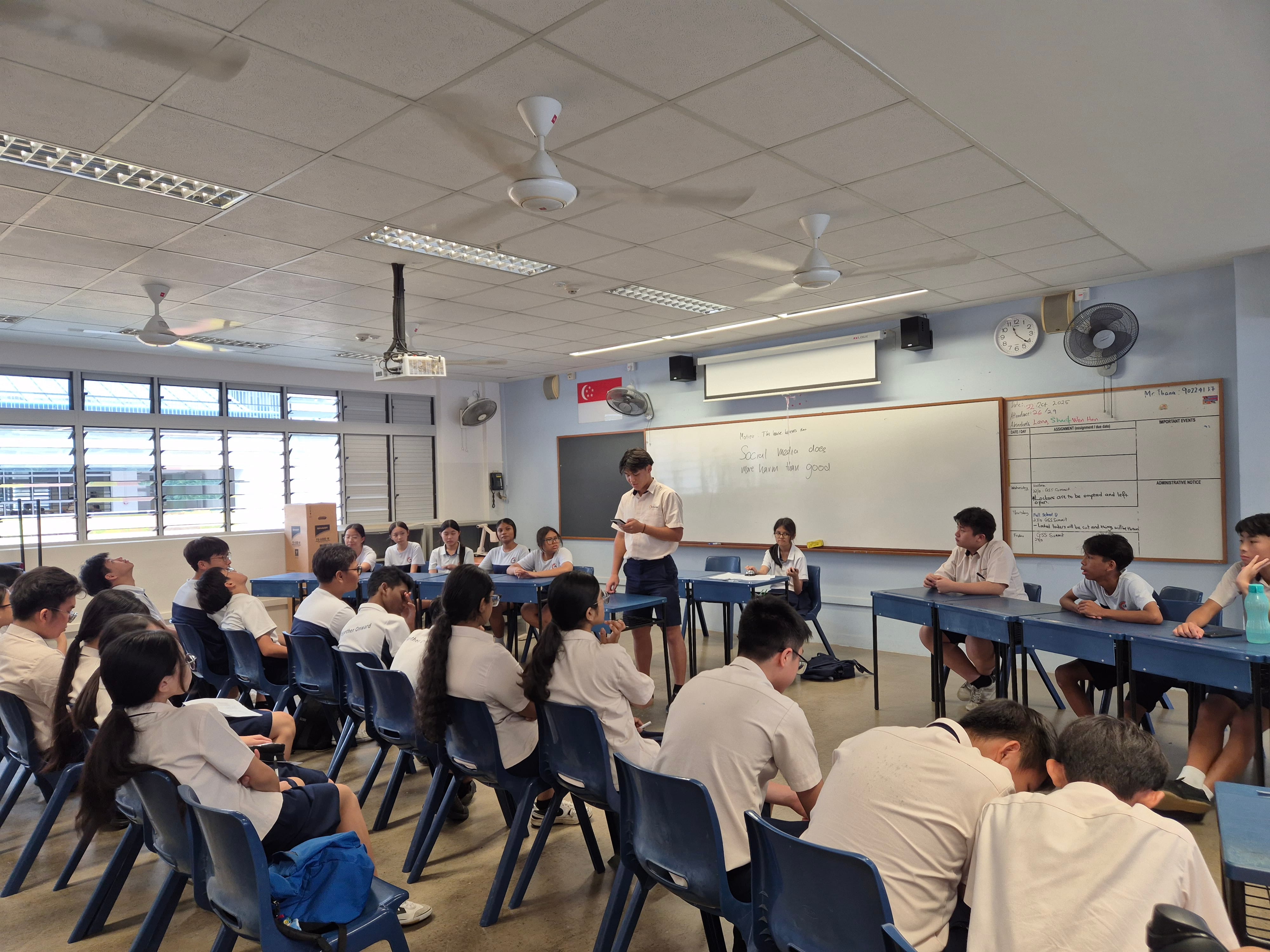 A student in uniform stands presenting to classmates seated in a school classroom with a whiteboard behind them.