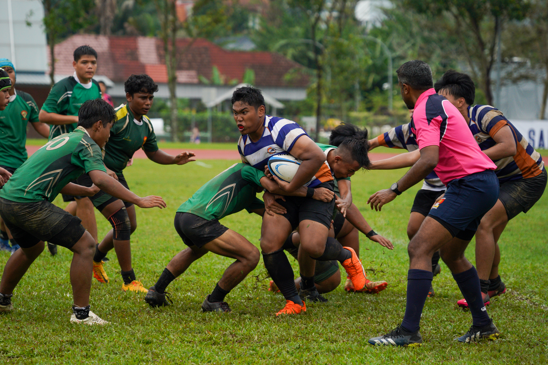 Rugby player in blue-and-white stripes runs with ball as green-clad opponents tackle on a muddy field.
