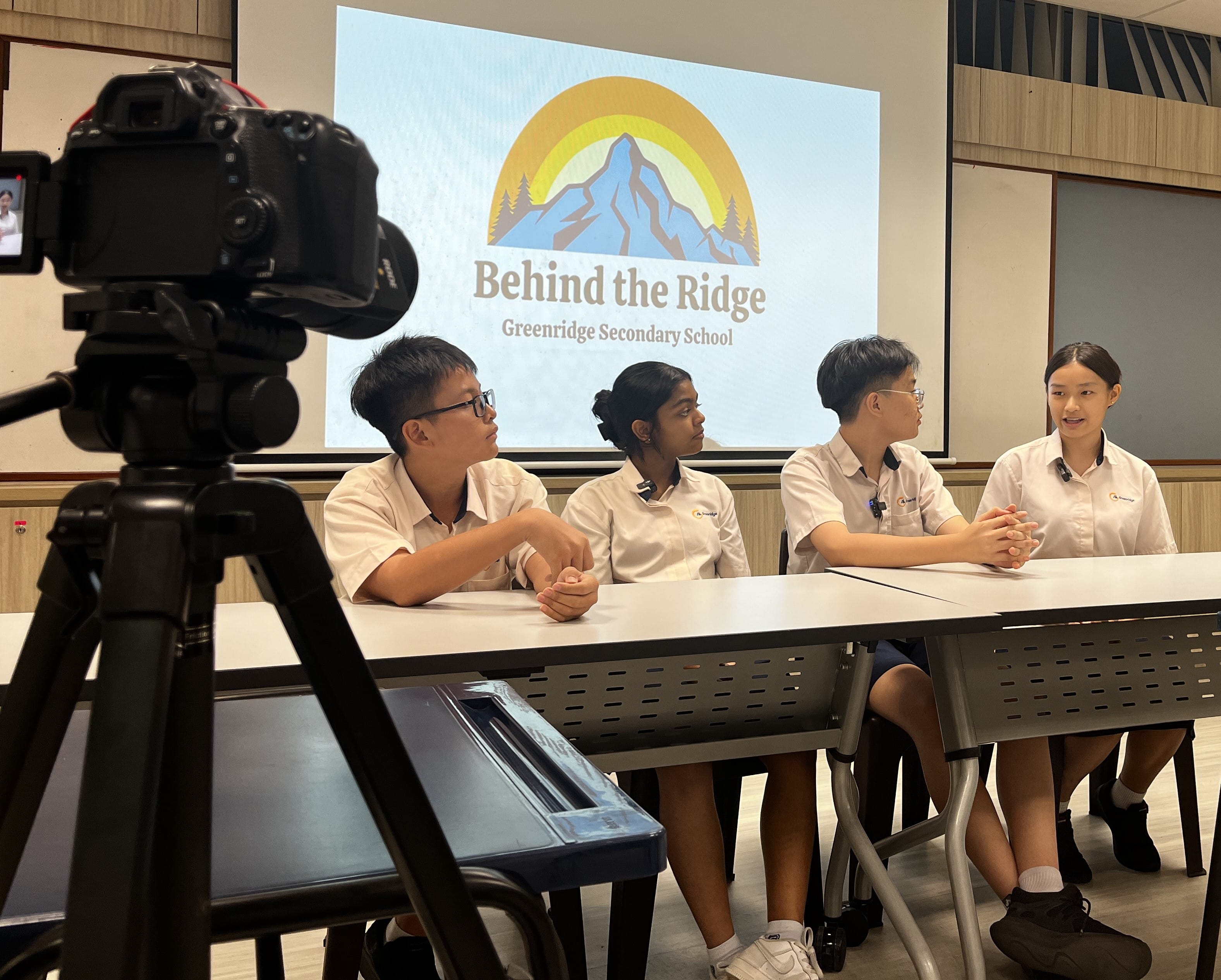 Four students in white uniforms sit at a table being filmed, with a Behind the Ridge Greenridge Secondary School screen behind them.
