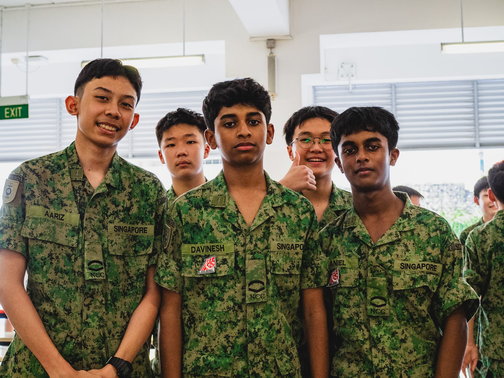 Five young male Singapore NCC cadets in green camouflage uniforms posing together indoors.