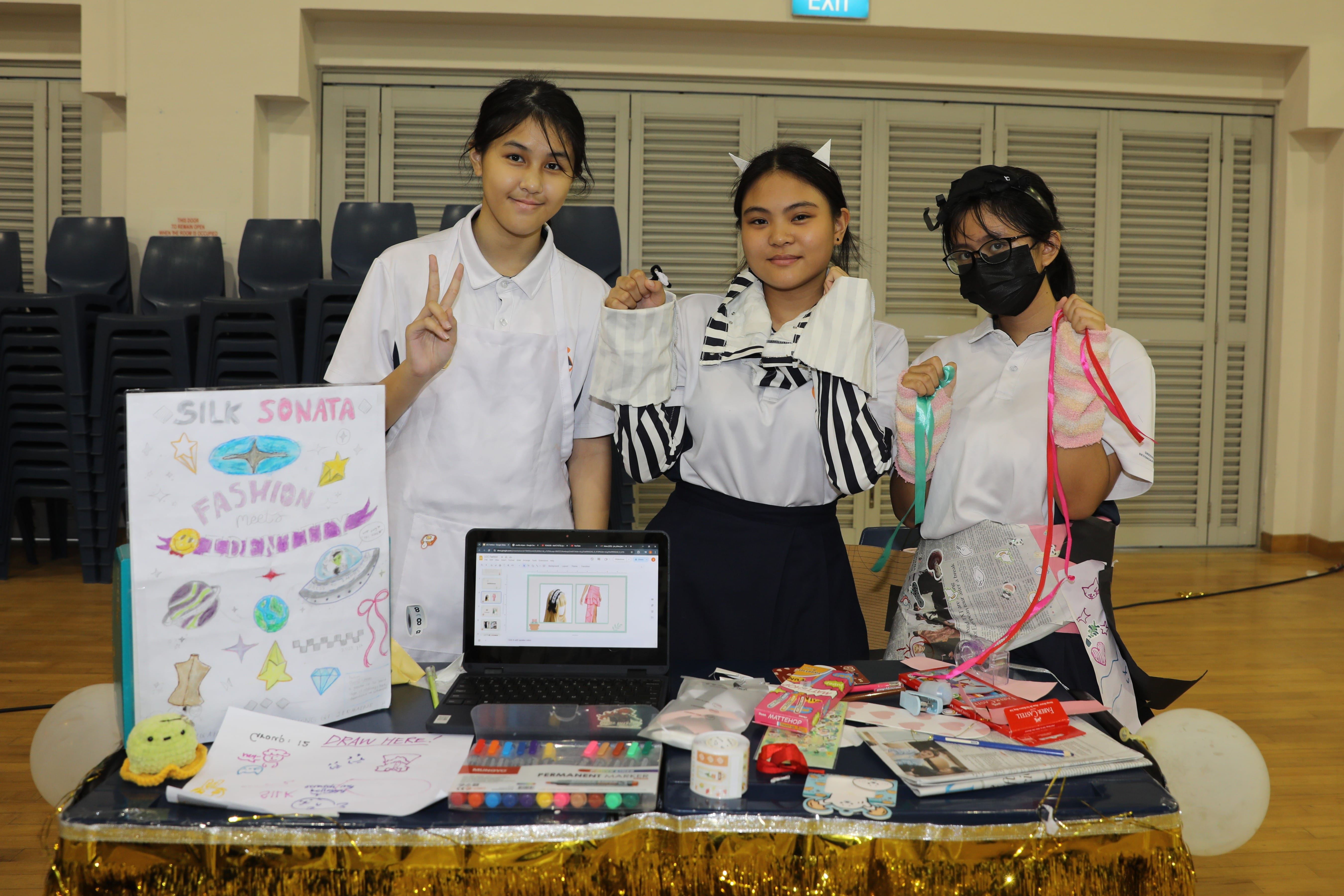 Three school girls stand at a table displaying a Silk Sonata Fashion project with a poster, laptop, and craft supplies.
