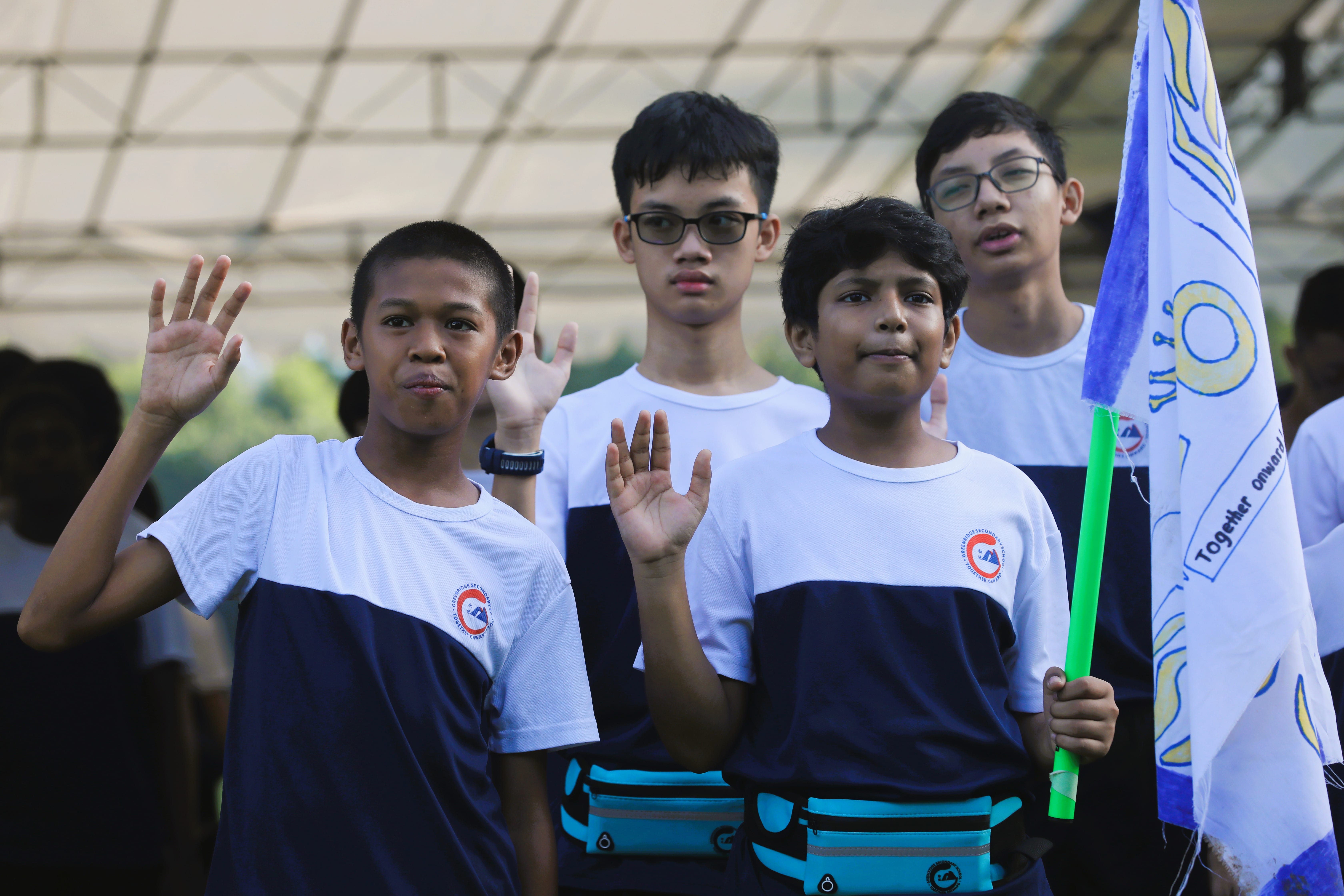 Four boys in matching white and navy school shirts raise their hands, one holding a blue flag with "Together Onward."