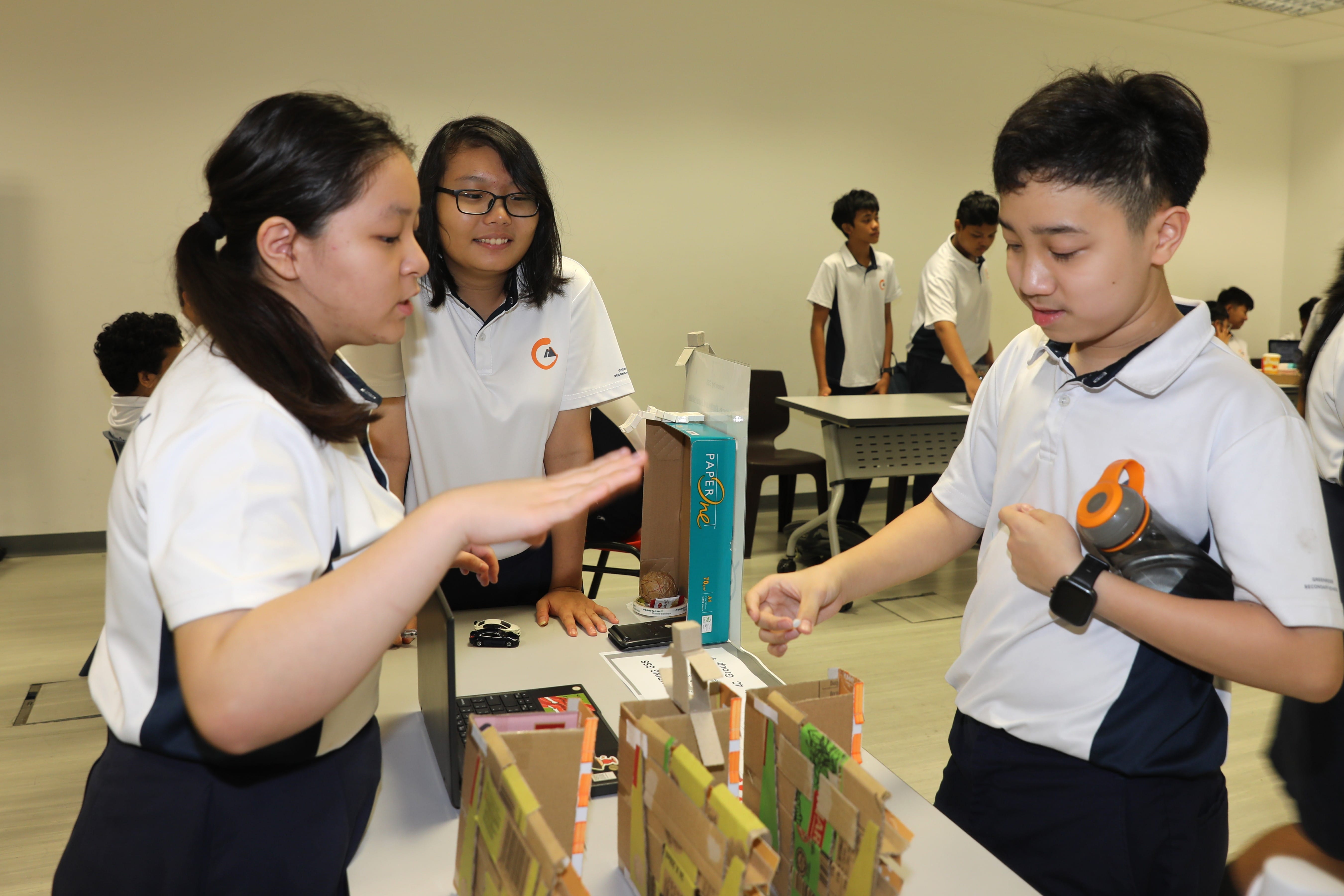 Three students in white uniforms work together on a cardboard model project at a table in a classroom.