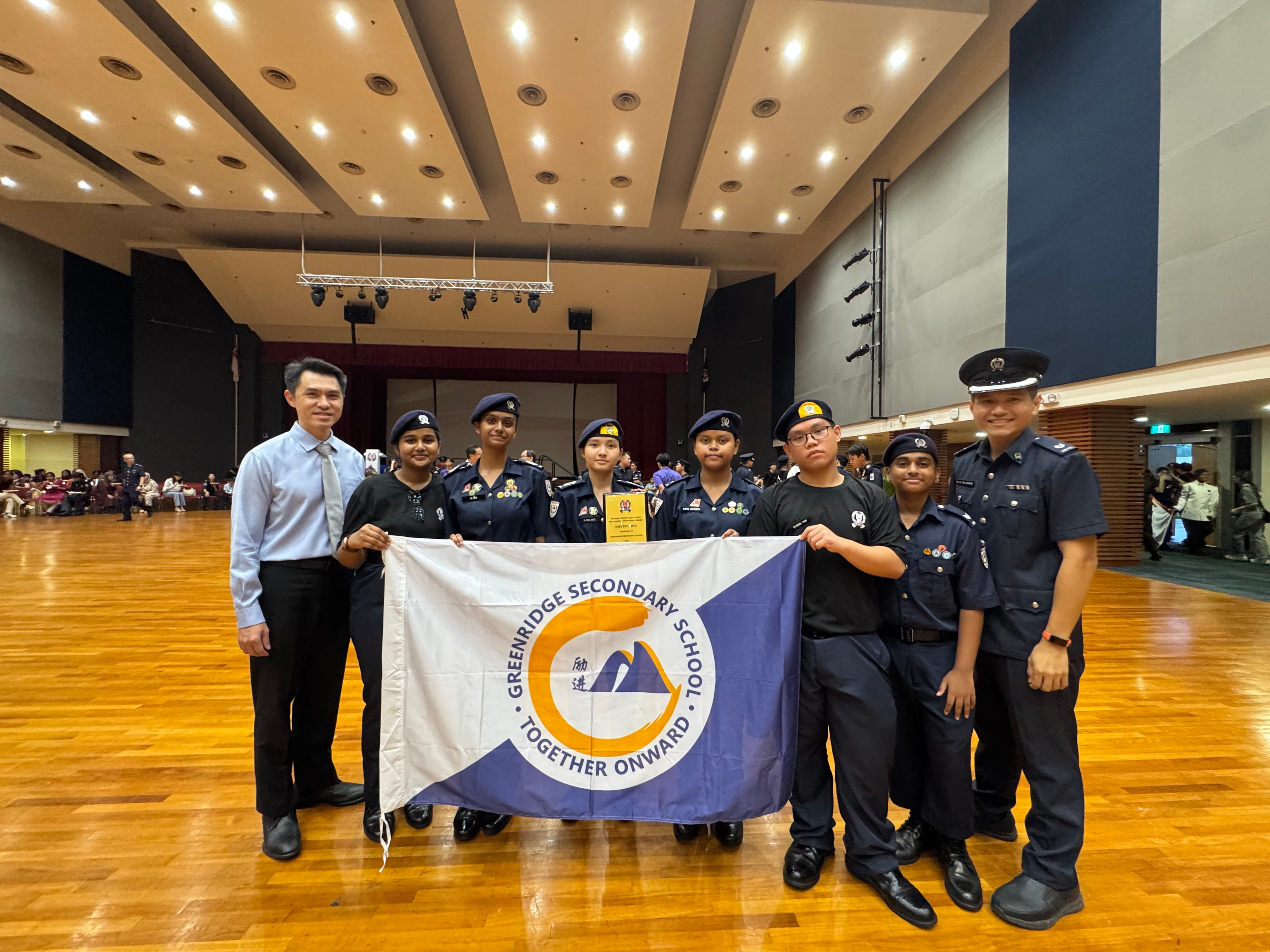 Group of uniformed students and adults holding a Greenridge Secondary School flag in a large hall.
