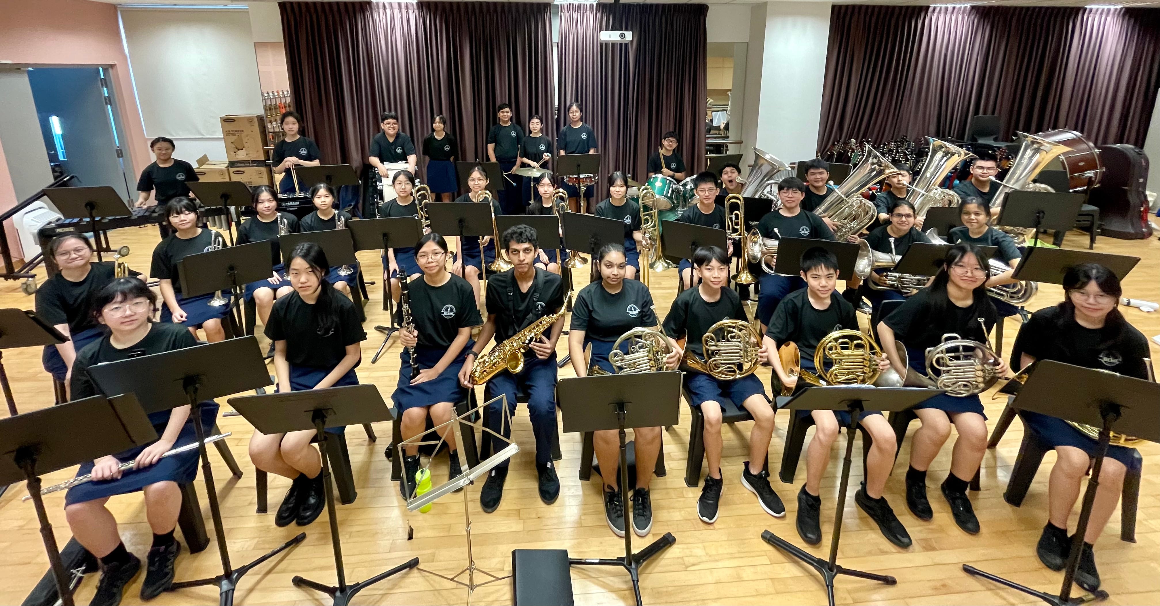 A large group of students in black uniforms sit with brass and woodwind instruments in a rehearsal room.