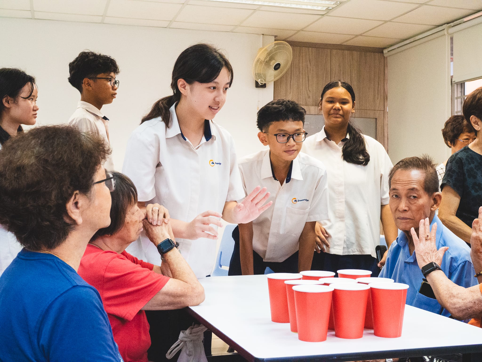 Students in white uniforms interact with elderly people at a table holding red plastic cups arranged in a triangle.