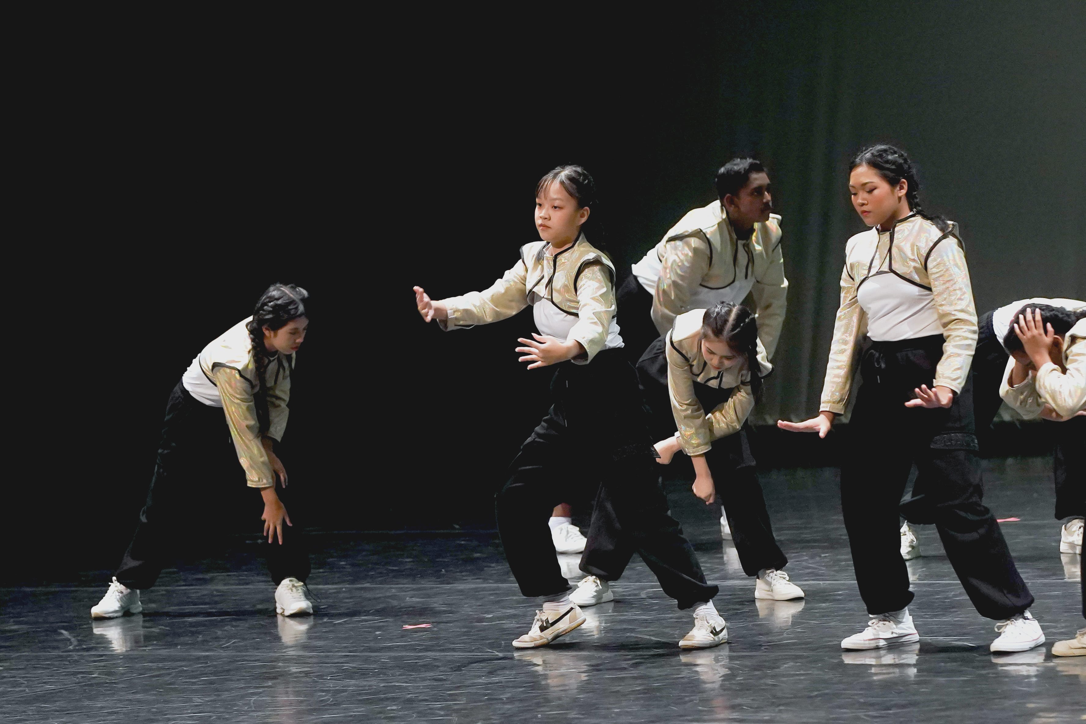A group of young dancers in matching gold and black outfits perform on a dark stage.