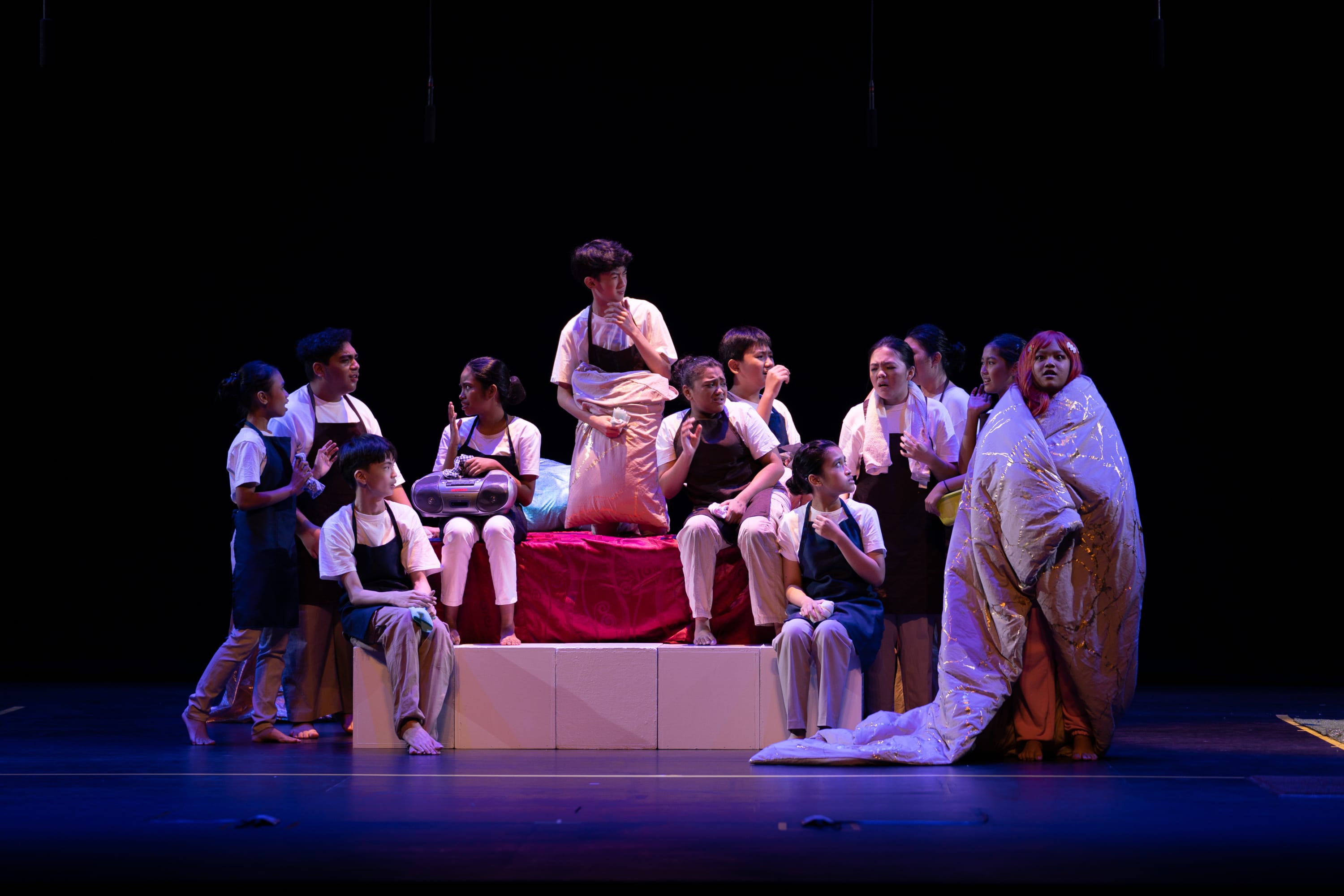 A group of young performers in white shirts and dark aprons gather around a red-draped platform on a dark stage.
