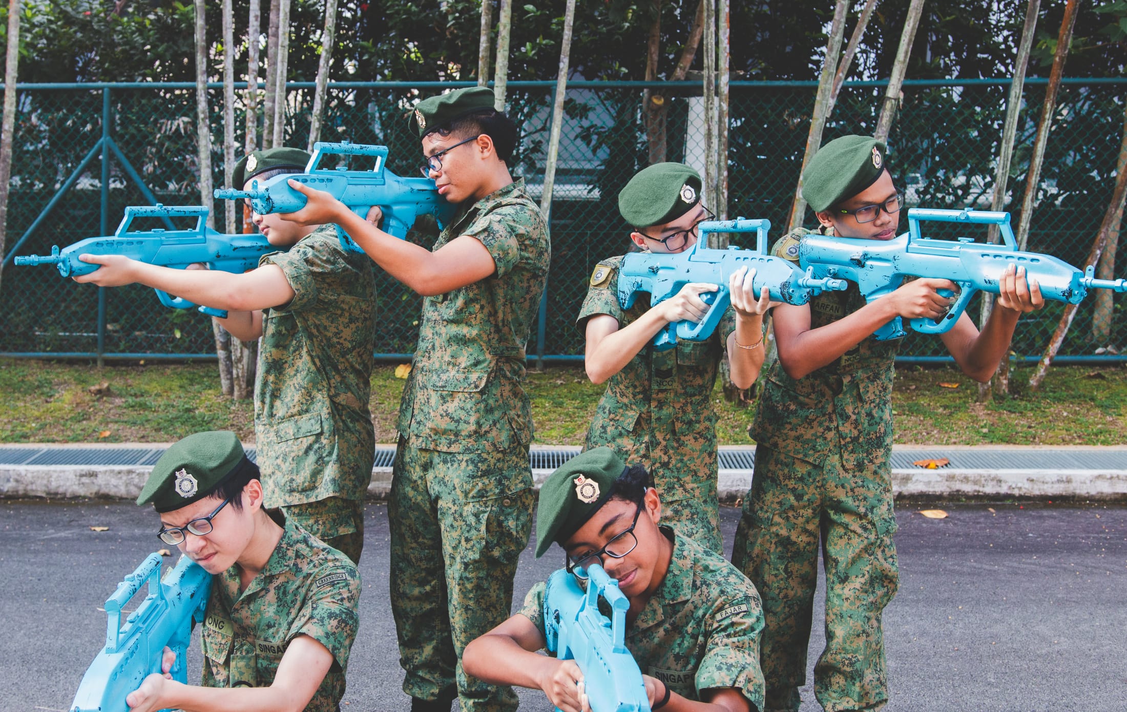 Six young cadets in camouflage uniforms and green berets aim blue training rifles outdoors.