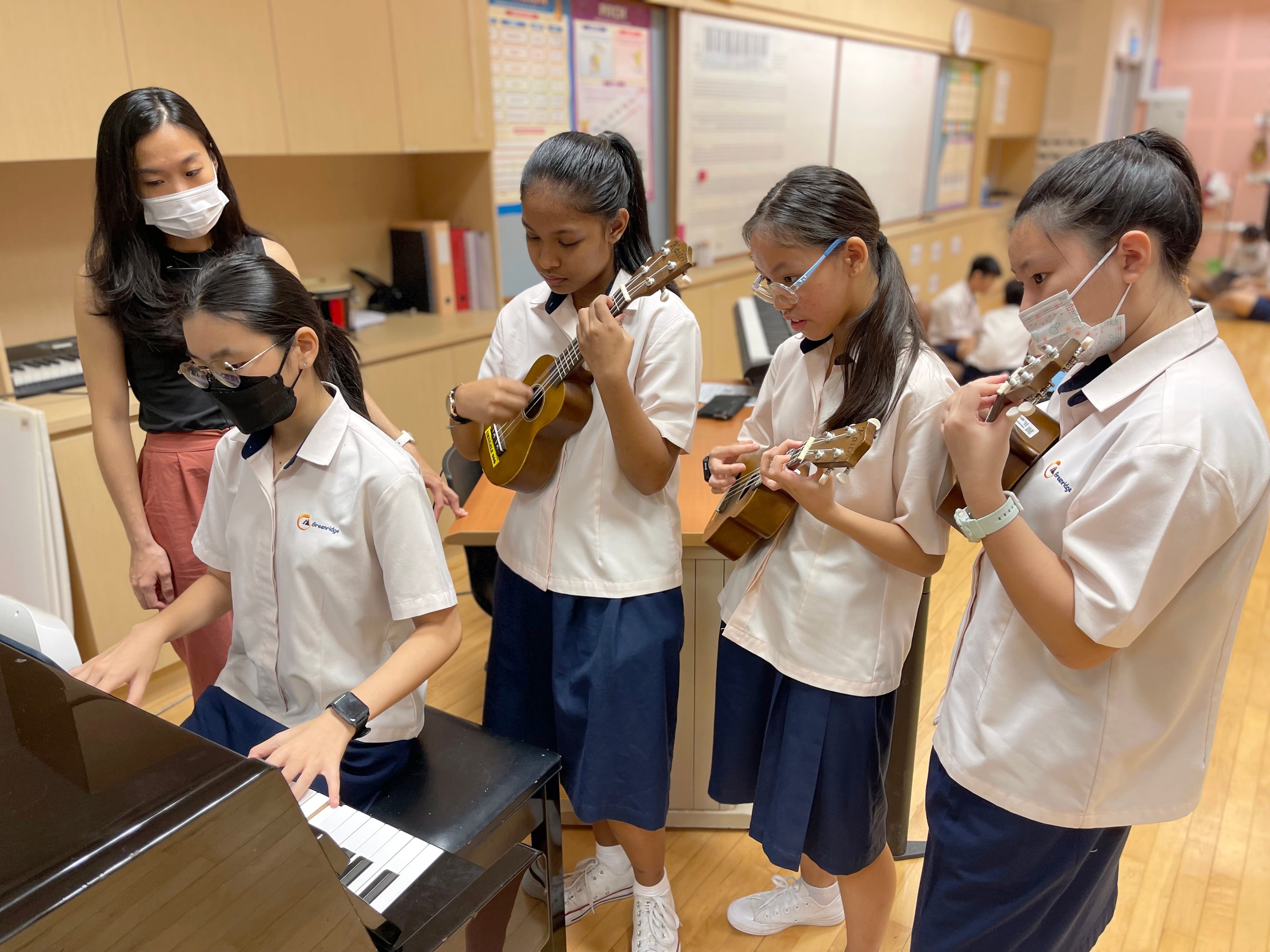 Teacher watches as one student plays piano and three students play ukuleles in a classroom.