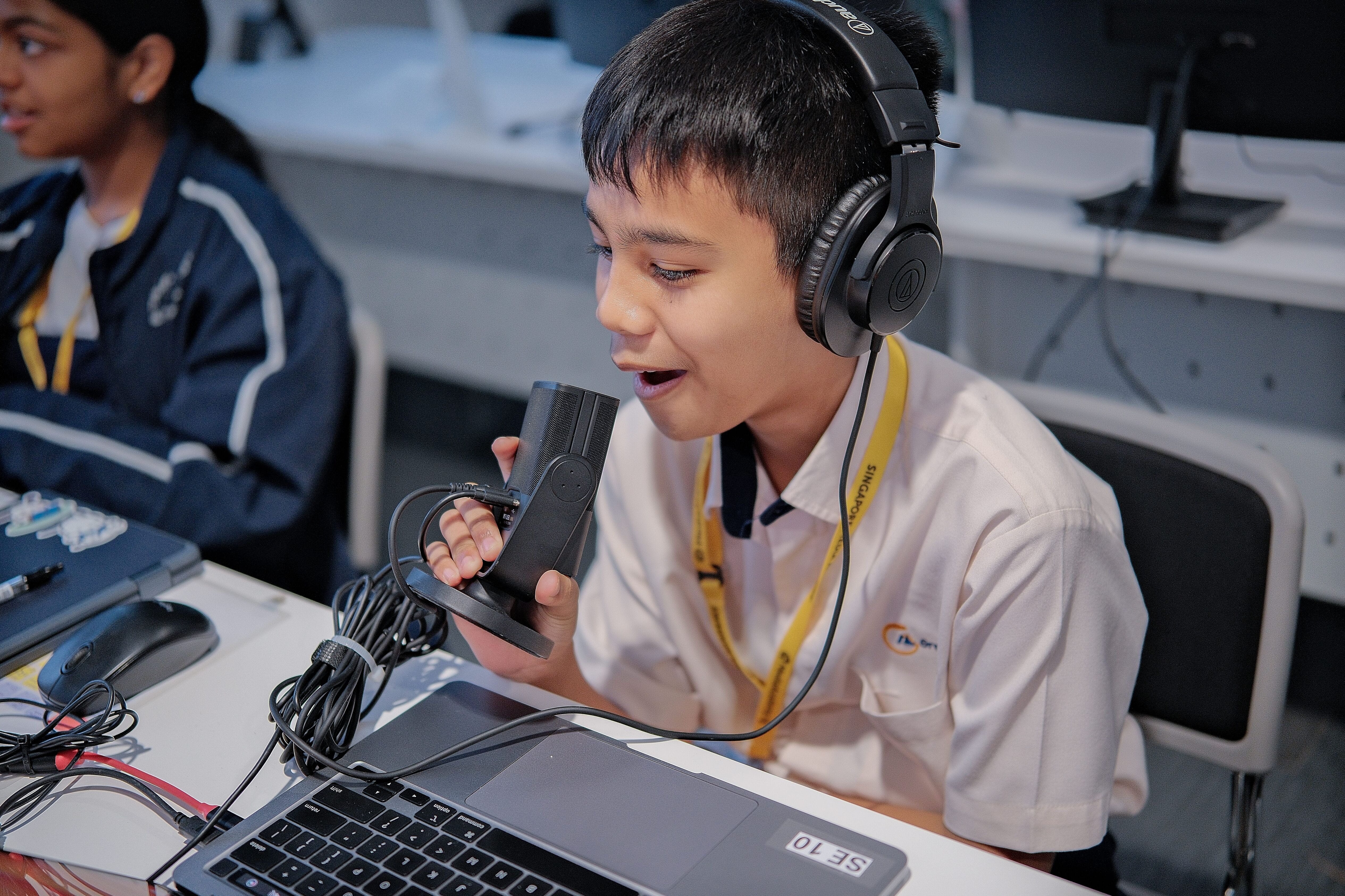 Student wearing headphones speaks into a handheld microphone at a desk with a laptop and cables.