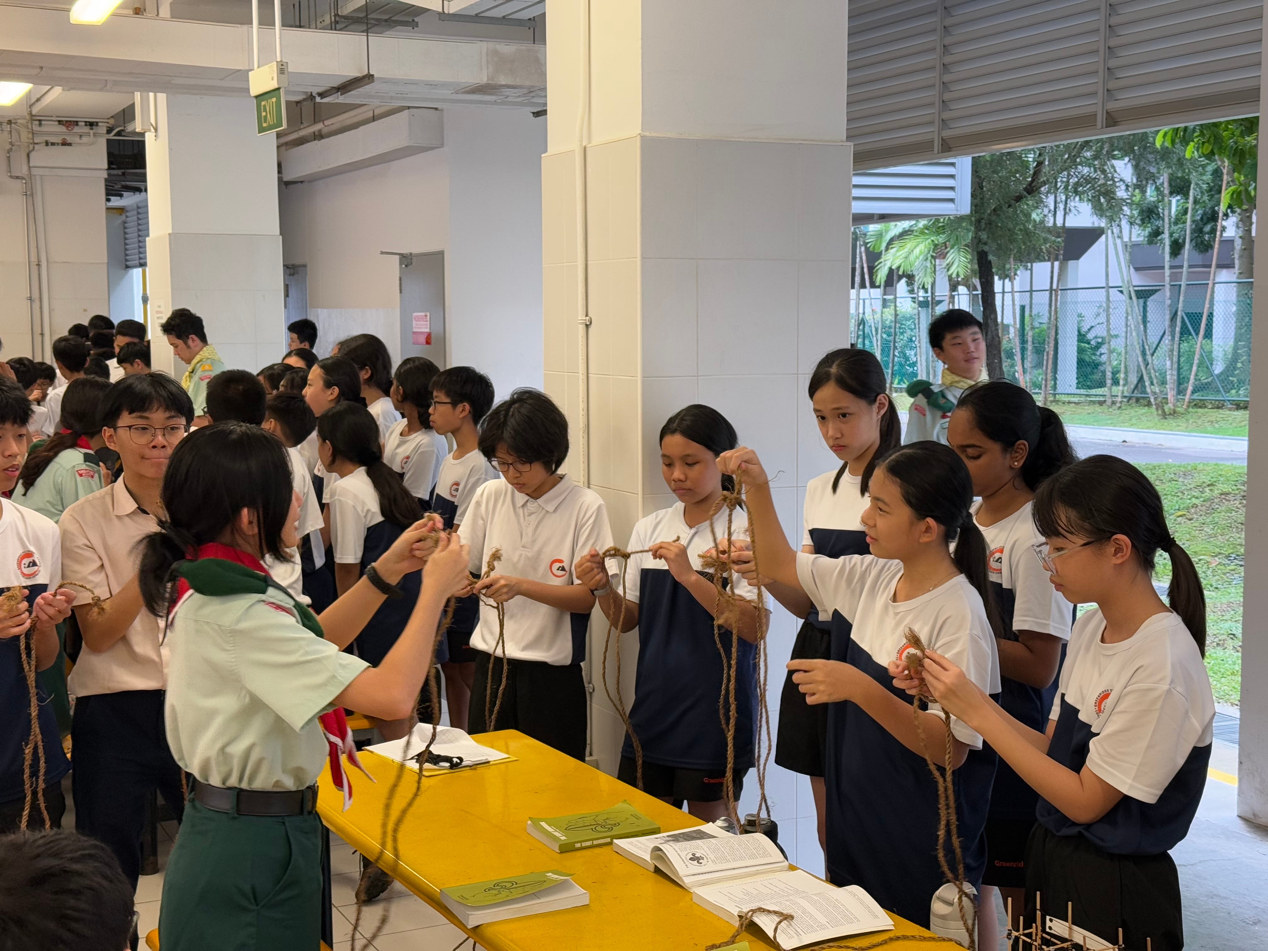 A scout in uniform teaches a group of students in school uniforms how to tie knots with rope indoors.