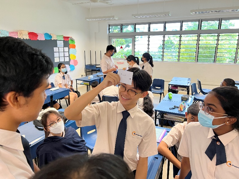 A smiling student in uniform holds up a card in a classroom while peers in masks look on.