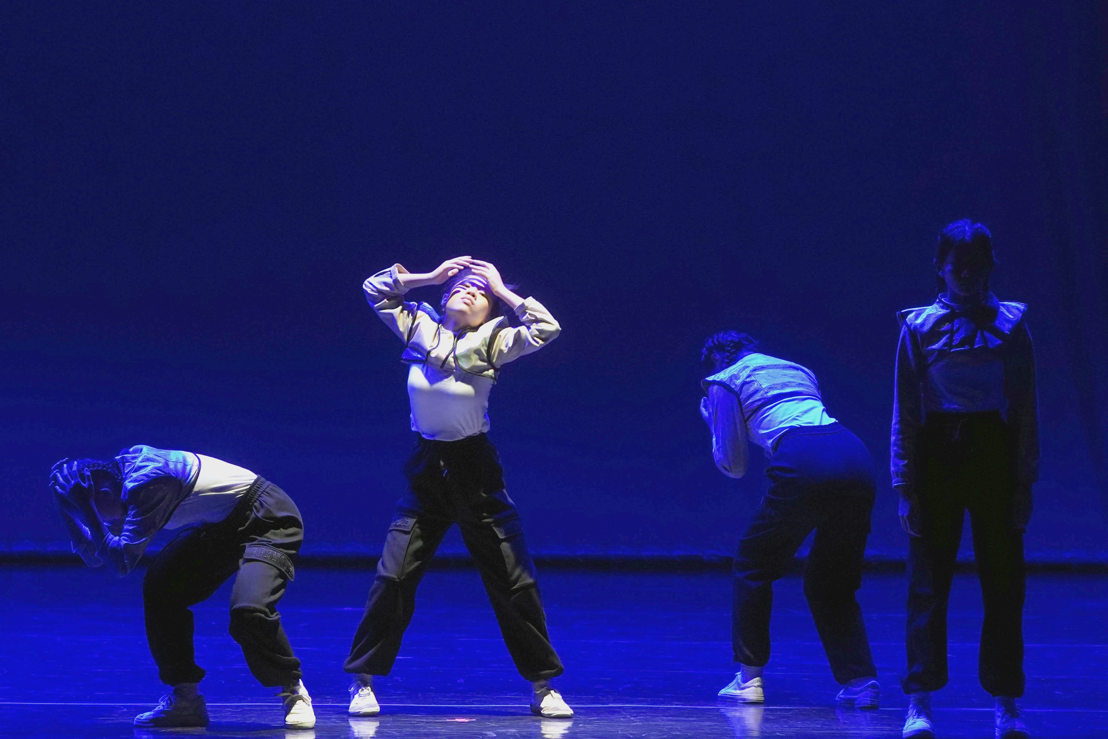 Four dancers perform on a blue-lit stage, one standing upright with hands on head while others crouch forward.