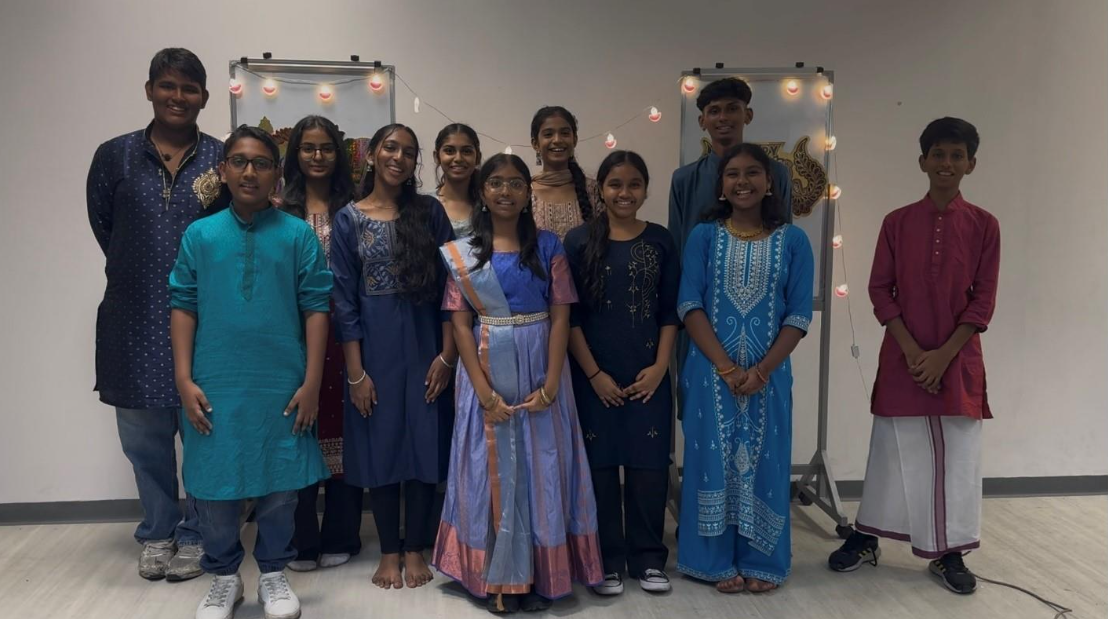 A group of young people wearing colorful traditional Indian attire pose together indoors near whiteboards with lights.