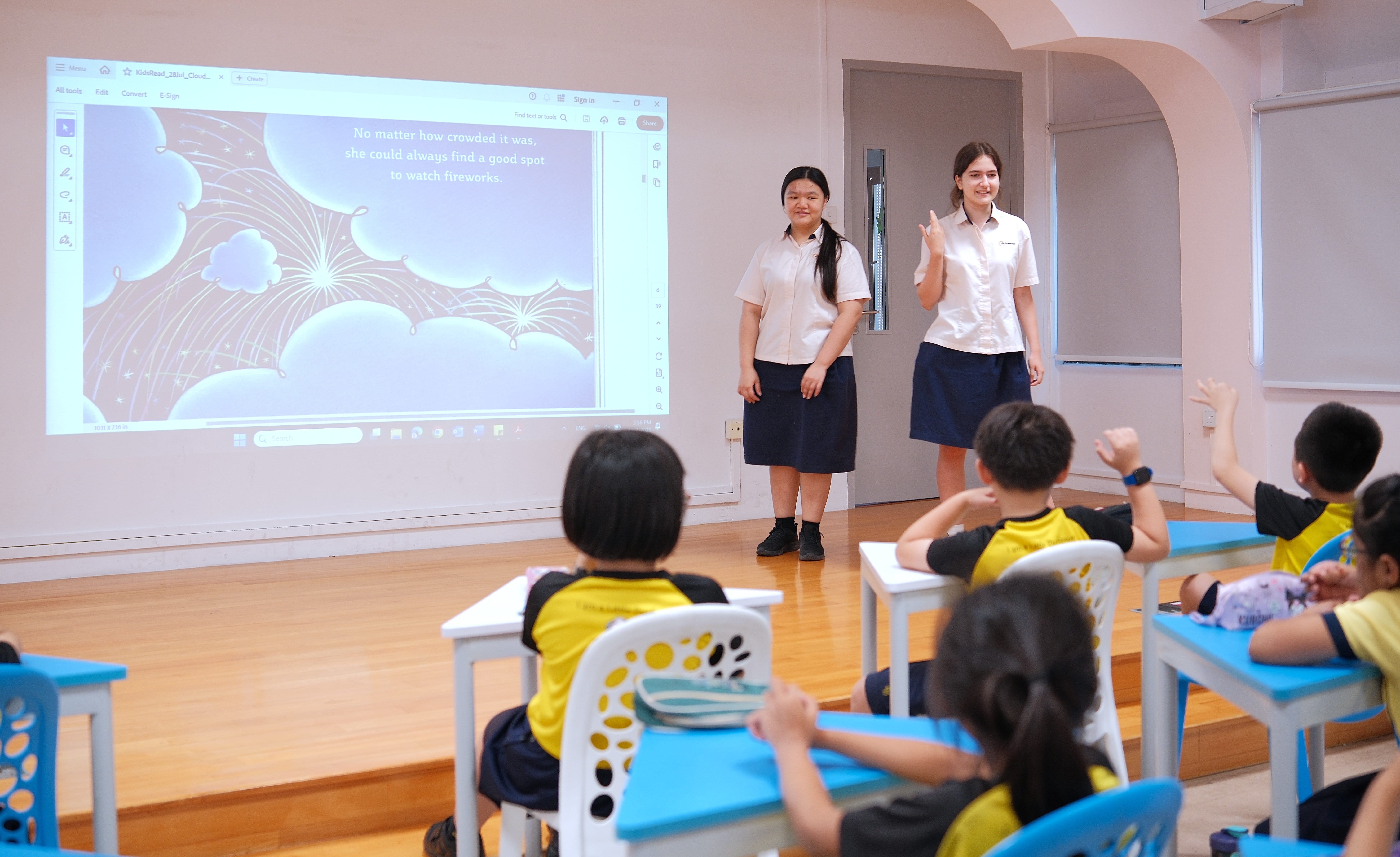Two students present beside a projected slide while children at desks raise their hands in a classroom.