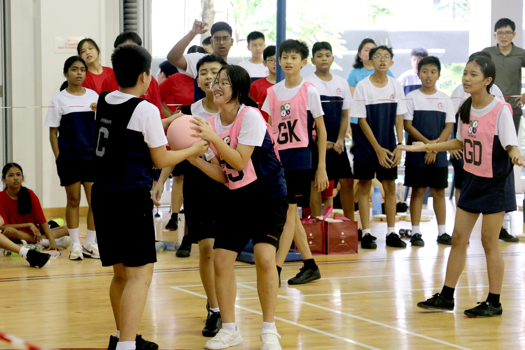 Students in pink and black uniforms play netball indoors, with one girl holding a pink ball while others watch.