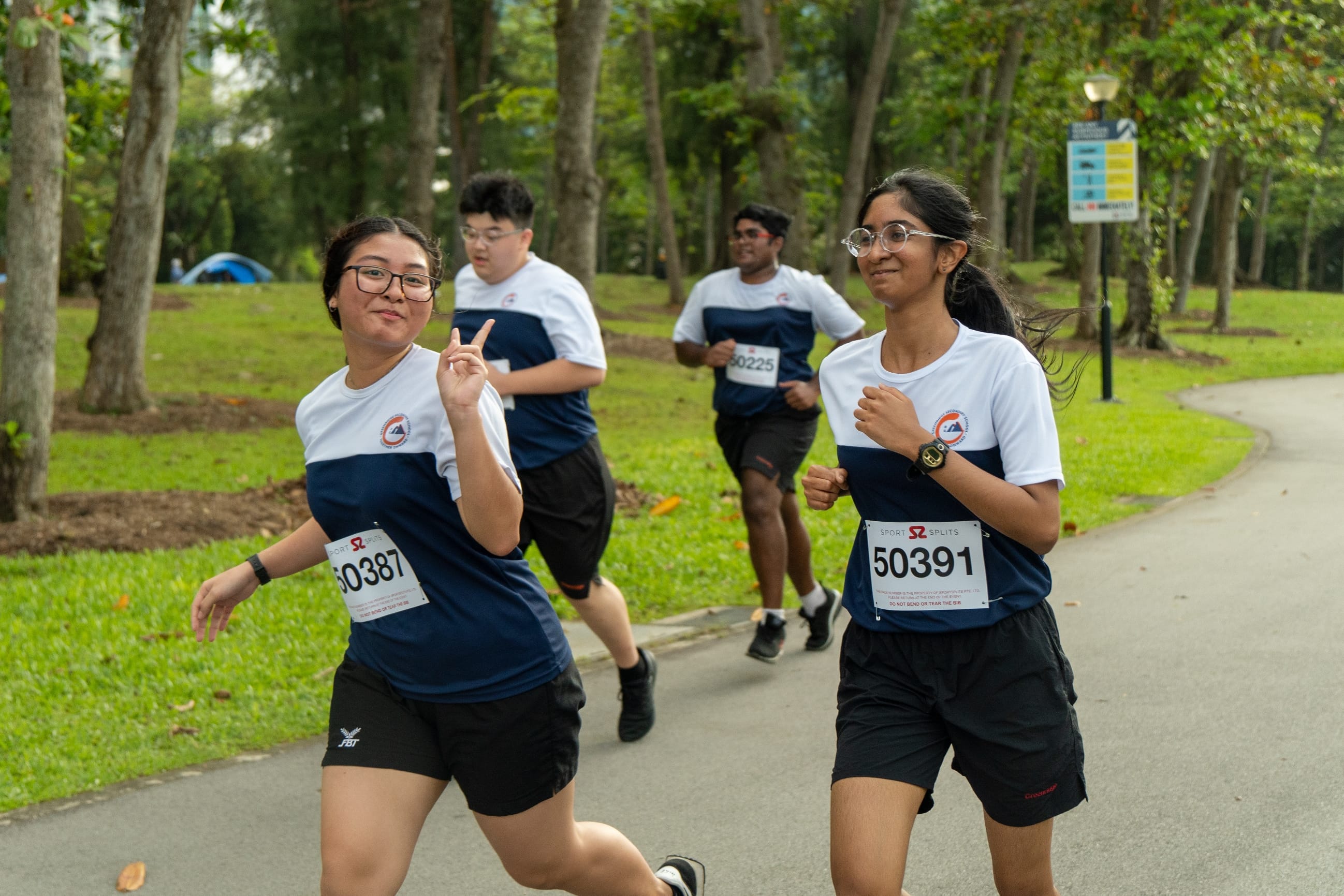 Four young runners in navy and white bibs race along a tree-lined park path, one flashing a peace sign.