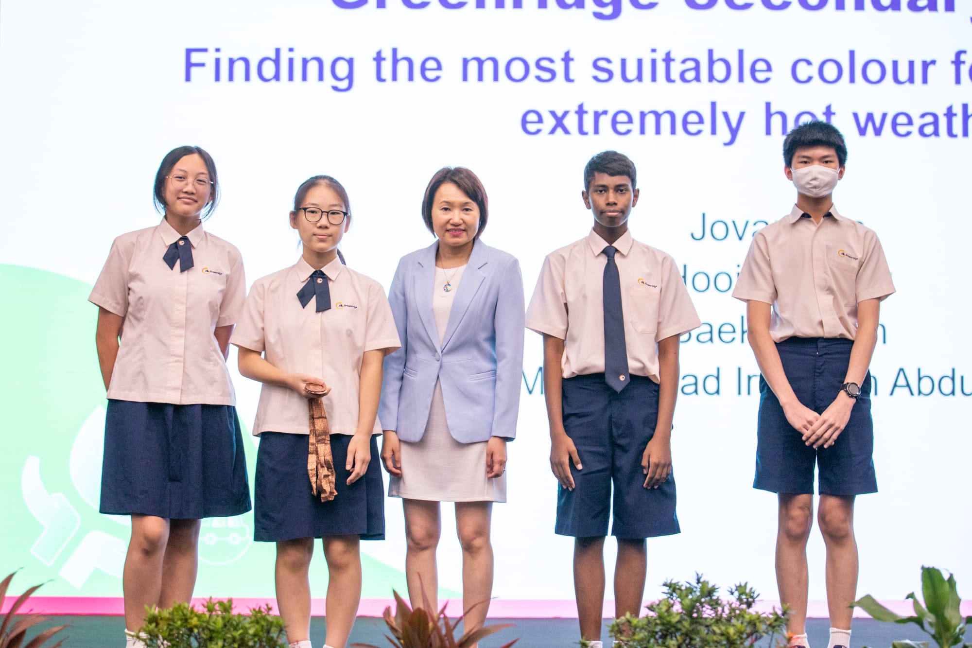 Five people in school uniforms and one adult in a blue blazer stand together on a stage in front of a presentation screen.
