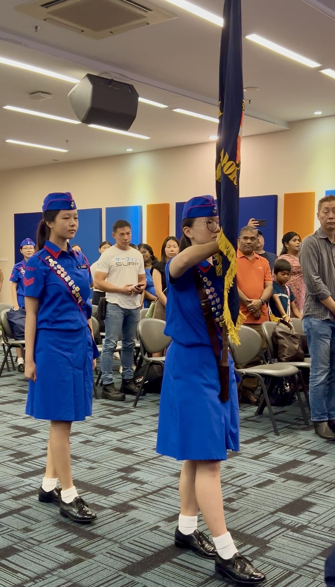 Two girls in blue uniforms and caps perform a flag ceremony indoors while spectators watch in the background.
