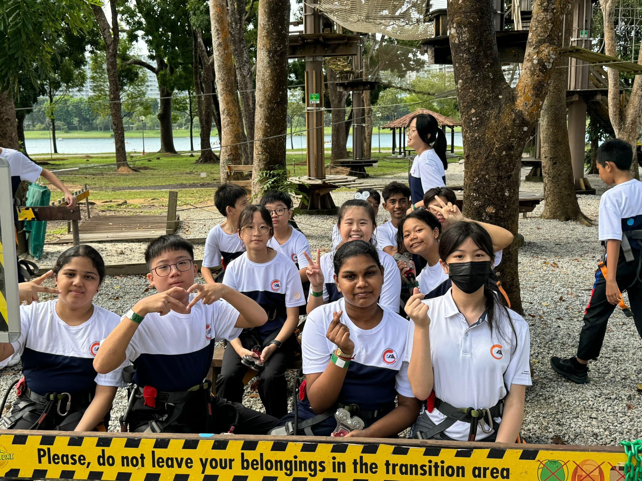 A group of students in white uniforms with harnesses pose smiling at an outdoor adventure park near trees.