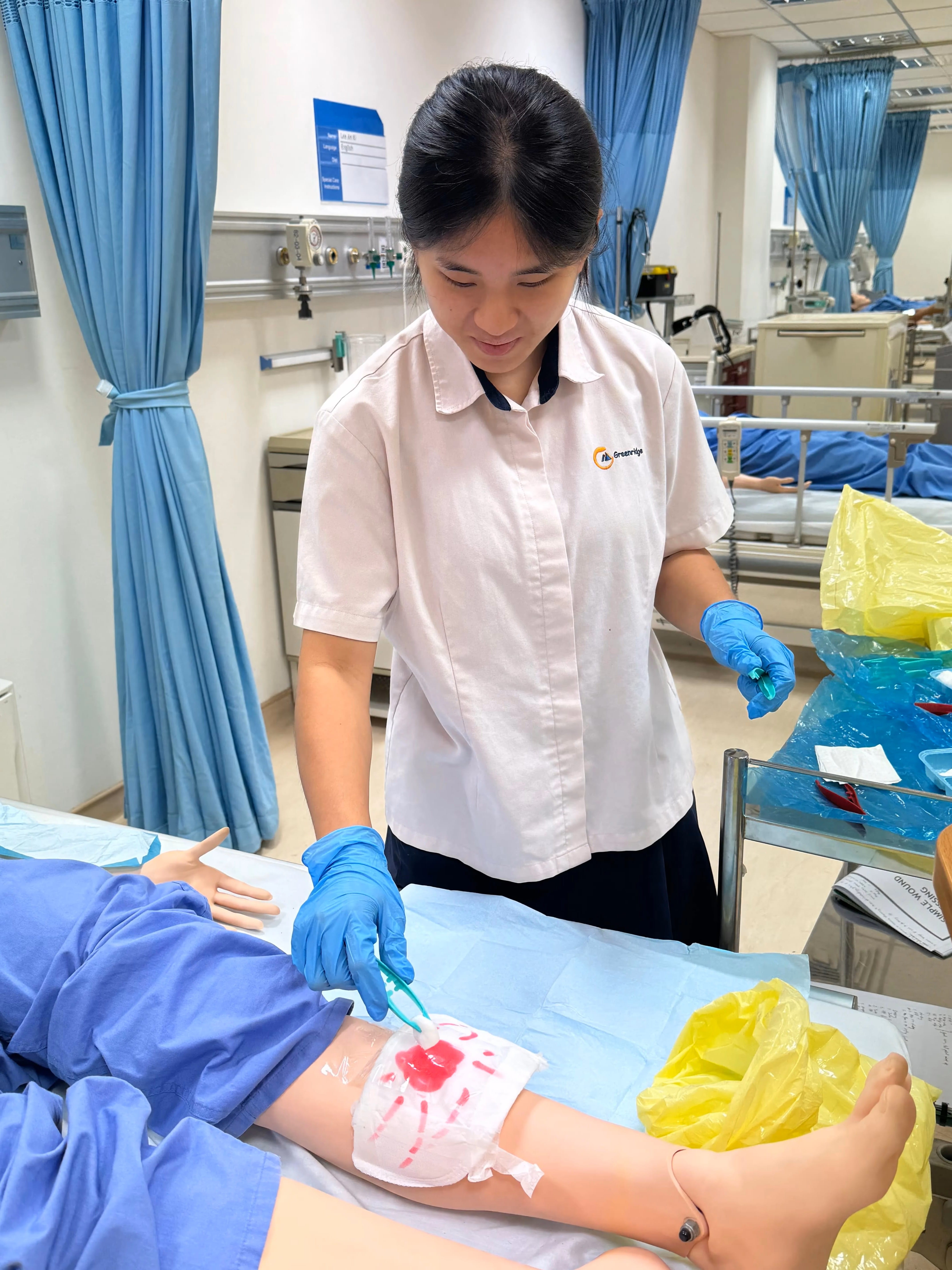 A woman in blue gloves practices wound care on a medical training mannequin leg in a simulation lab.