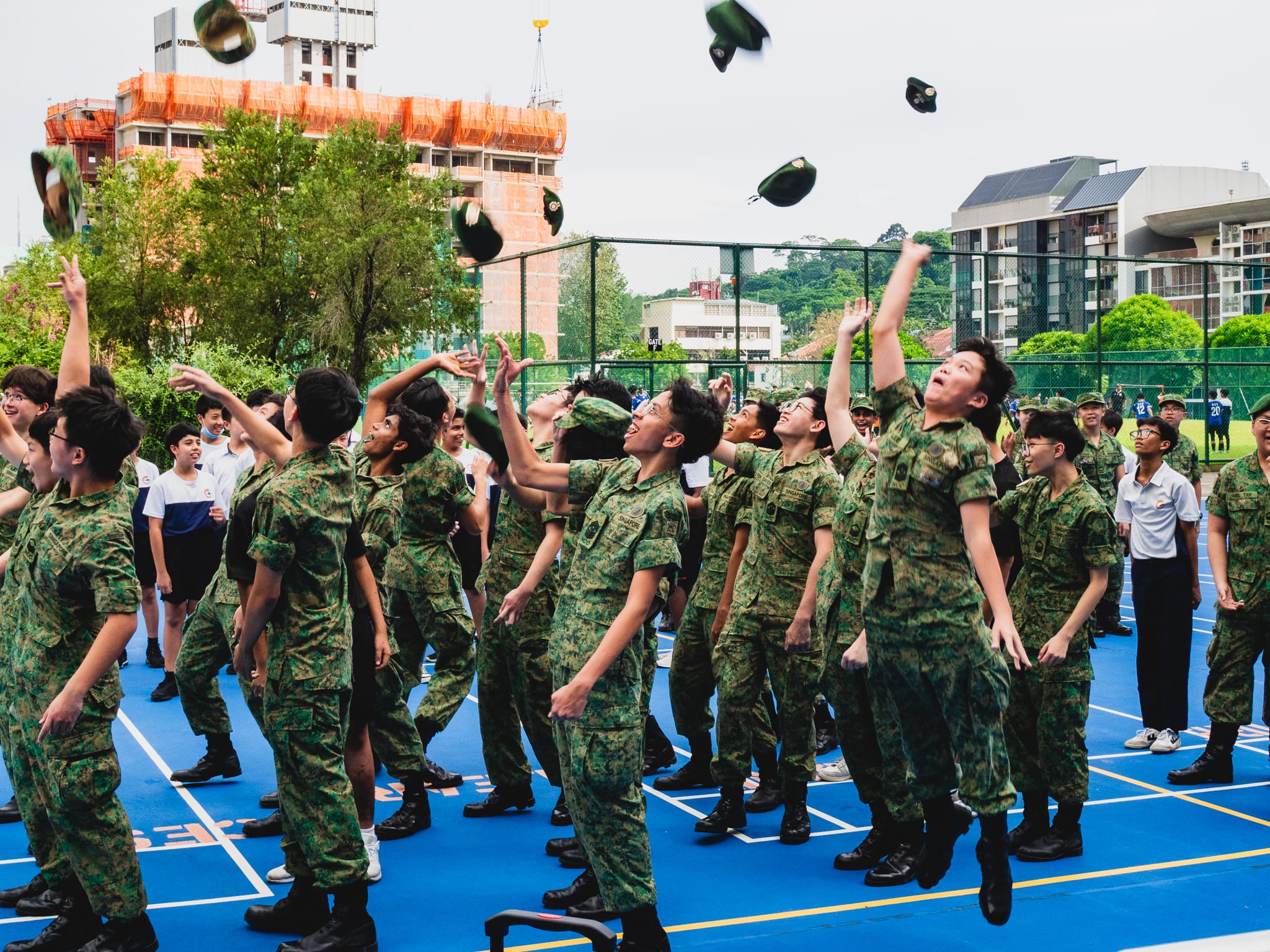 Young soldiers in camouflage uniforms joyfully toss their green caps into the air on a blue outdoor court.