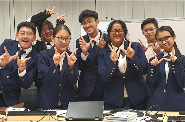 Eight students in navy blazers pose smiling, making hand signs, with a laptop on the table in front of them.