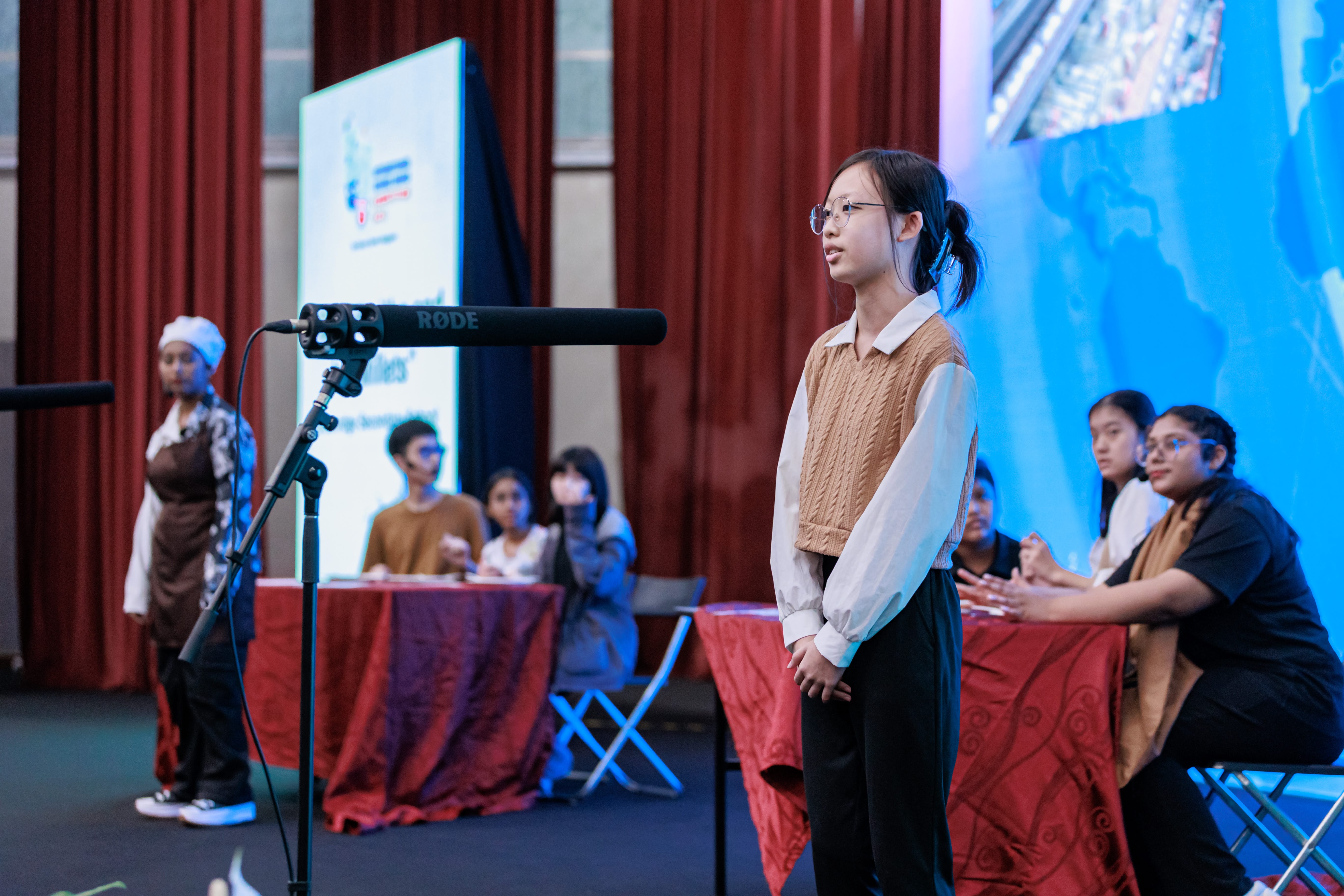 Girl stands by a boom microphone onstage as judges sit at tables; red curtains and blue screen behind.