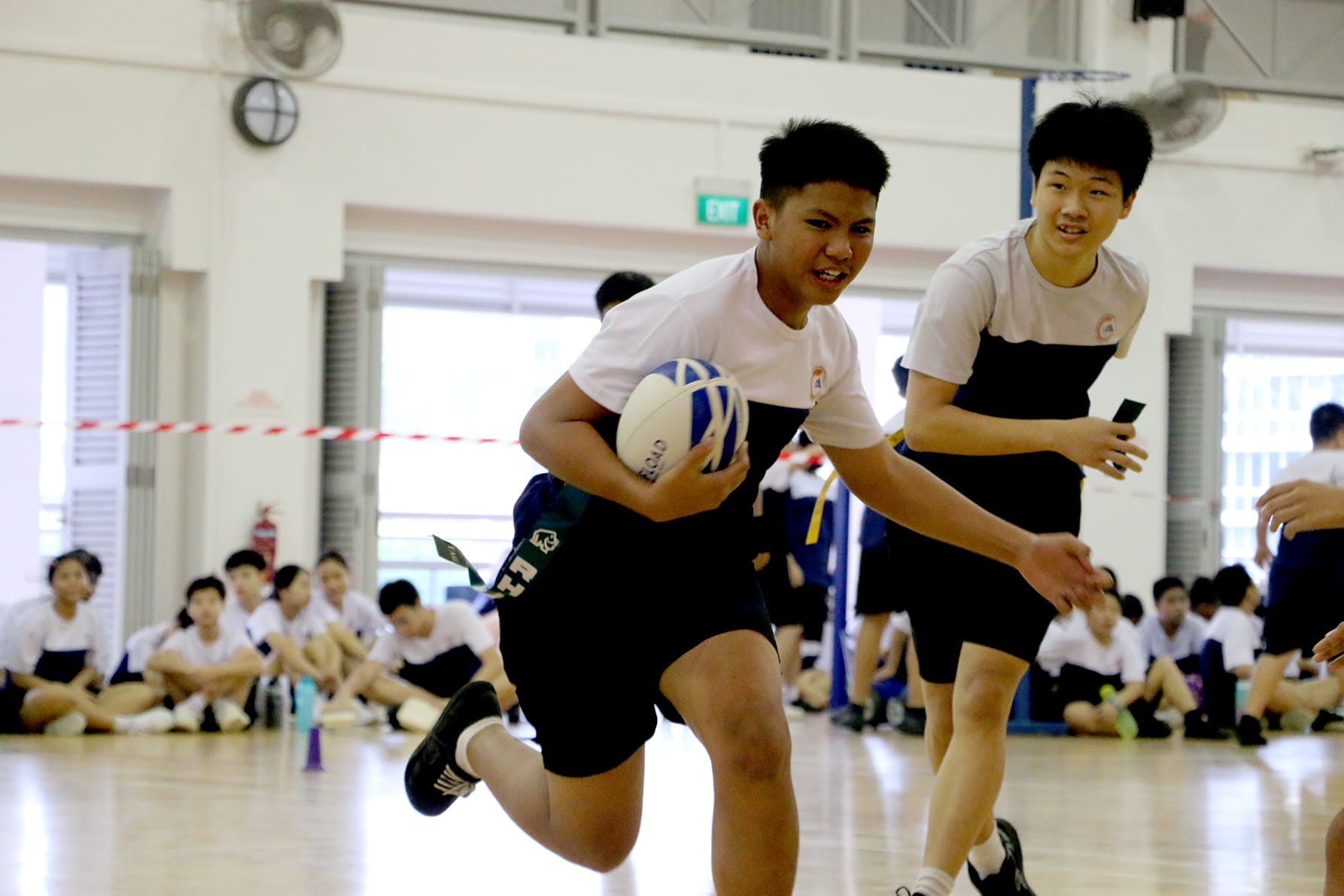 Two teenage boys in white shirts run with a rugby ball on an indoor court, with students watching in the background.