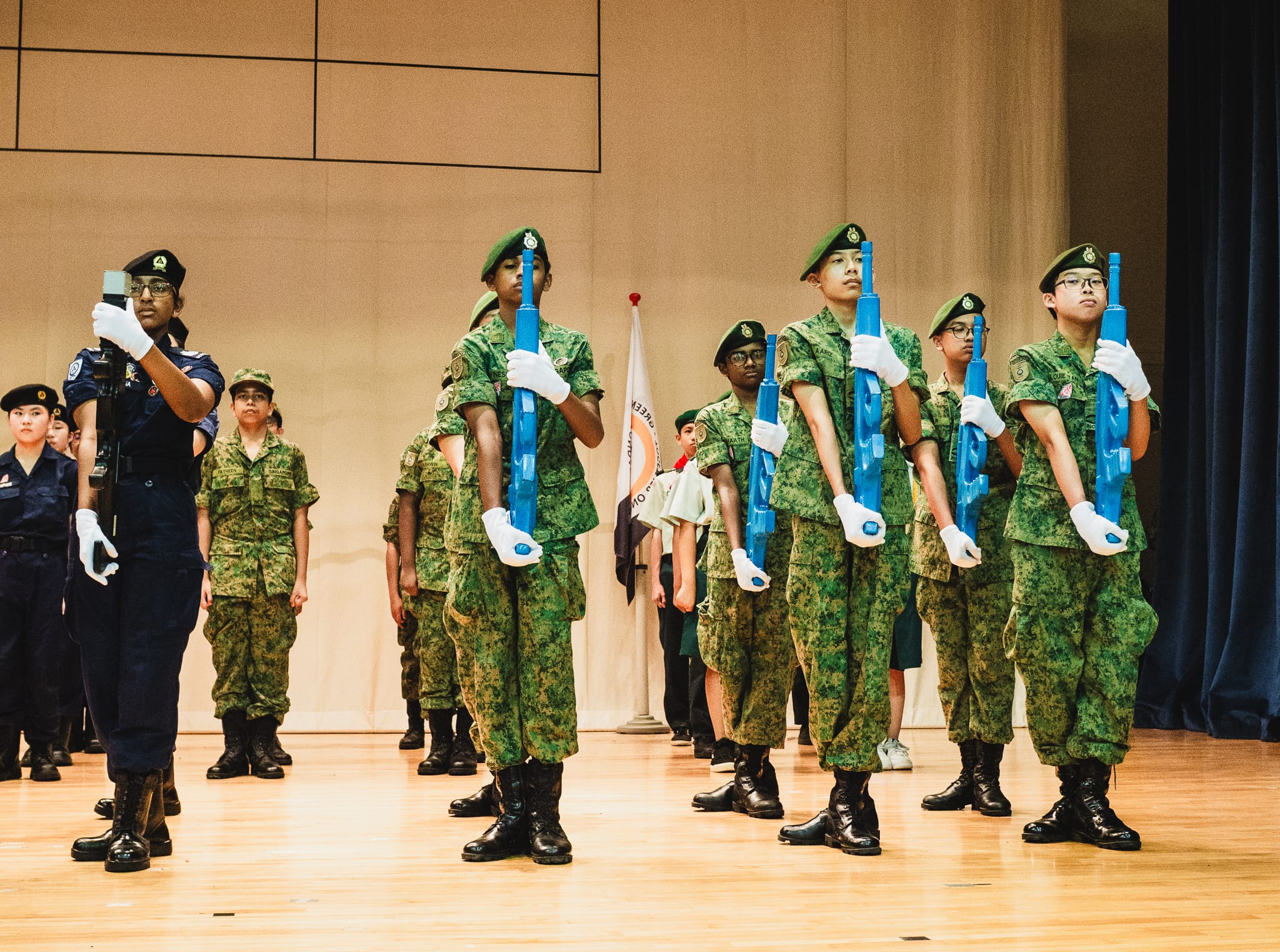 Young cadets in green camouflage uniforms hold blue rifles during a formal drill ceremony on a wooden stage.