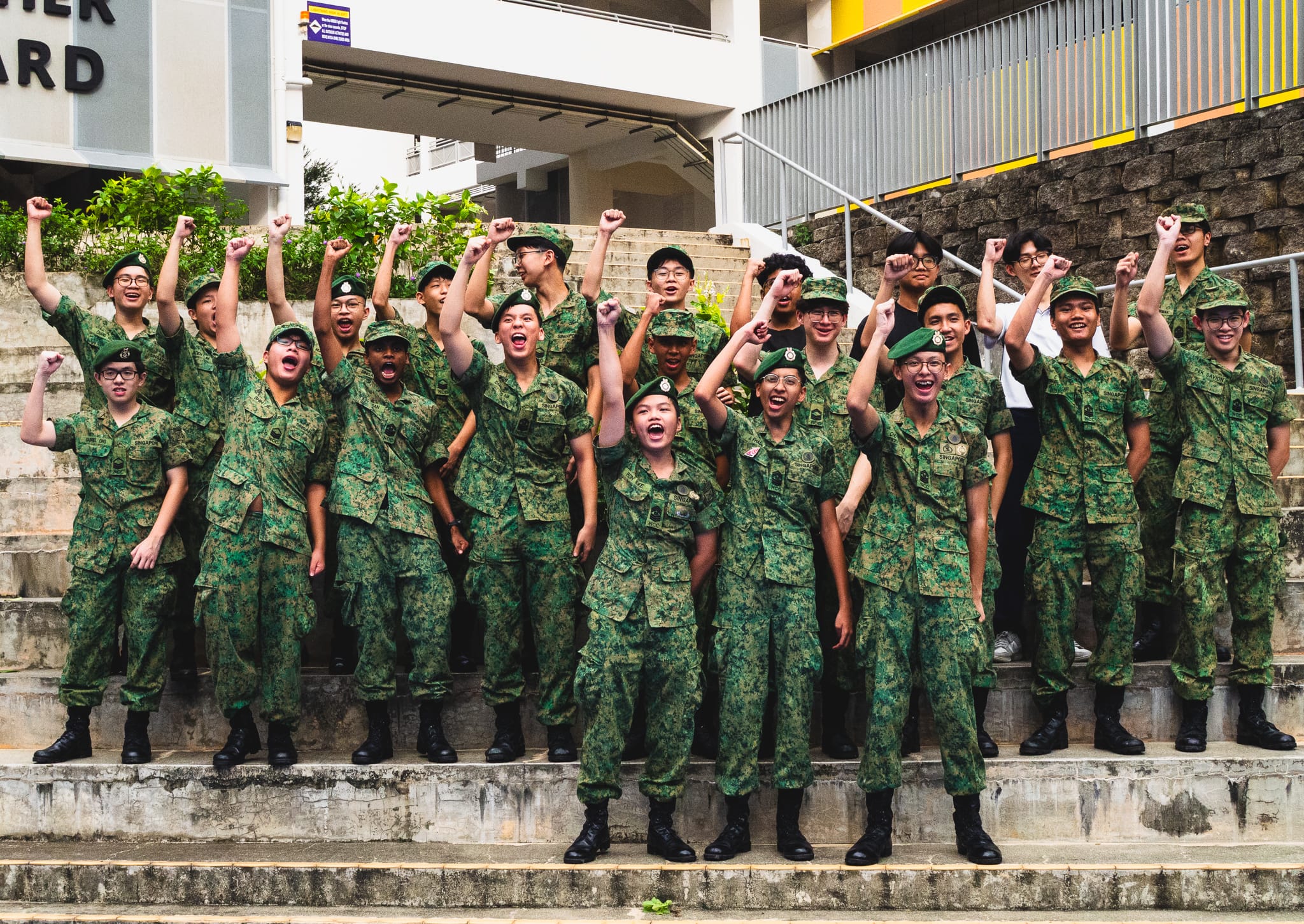 A large group of young people in green camouflage uniforms cheer with raised fists on outdoor steps.