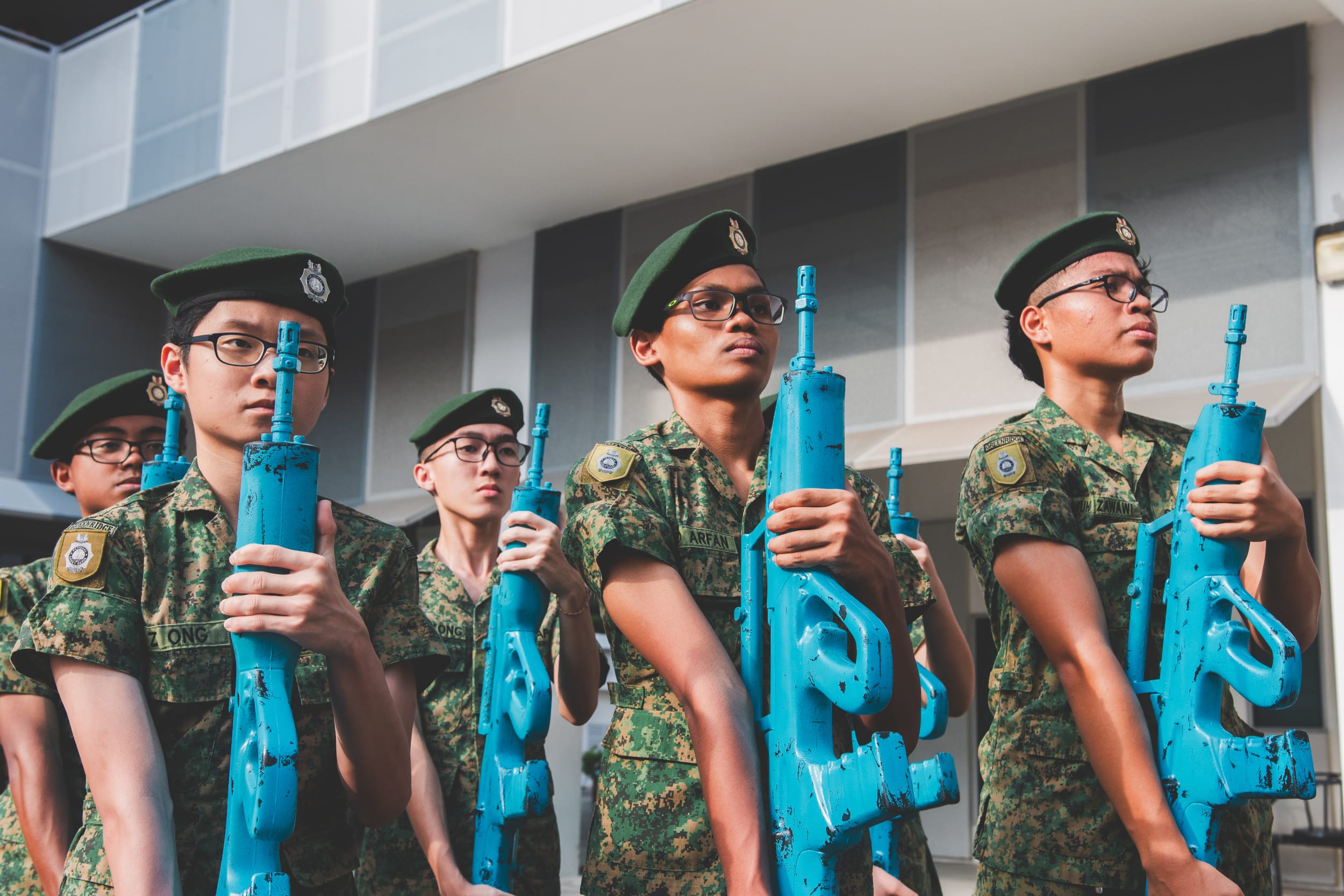 Young cadets in camouflage uniforms and green berets stand in formation holding blue training rifles outdoors.