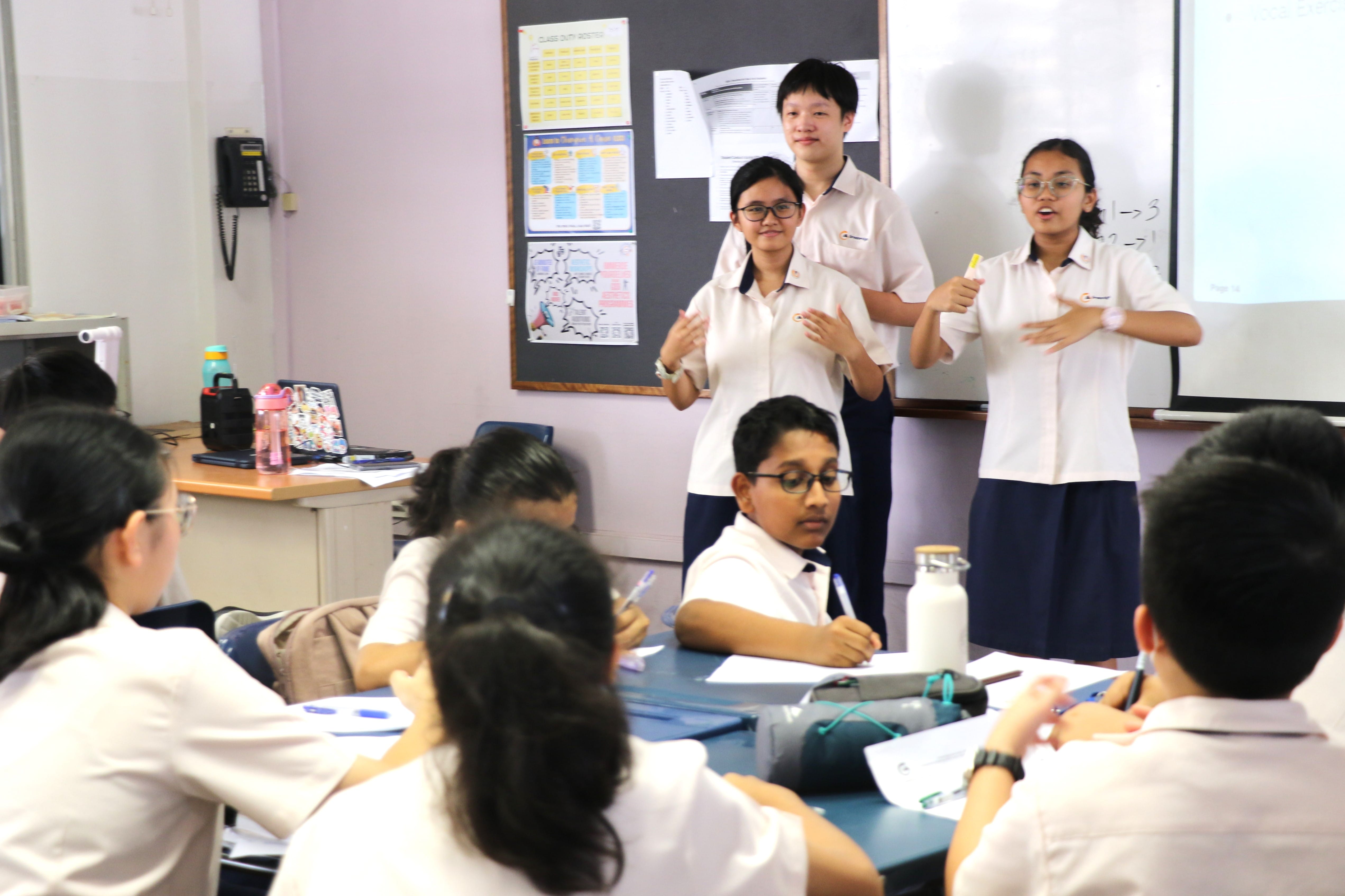 Three students in white uniforms present at the front of a classroom while seated classmates listen and take notes.
