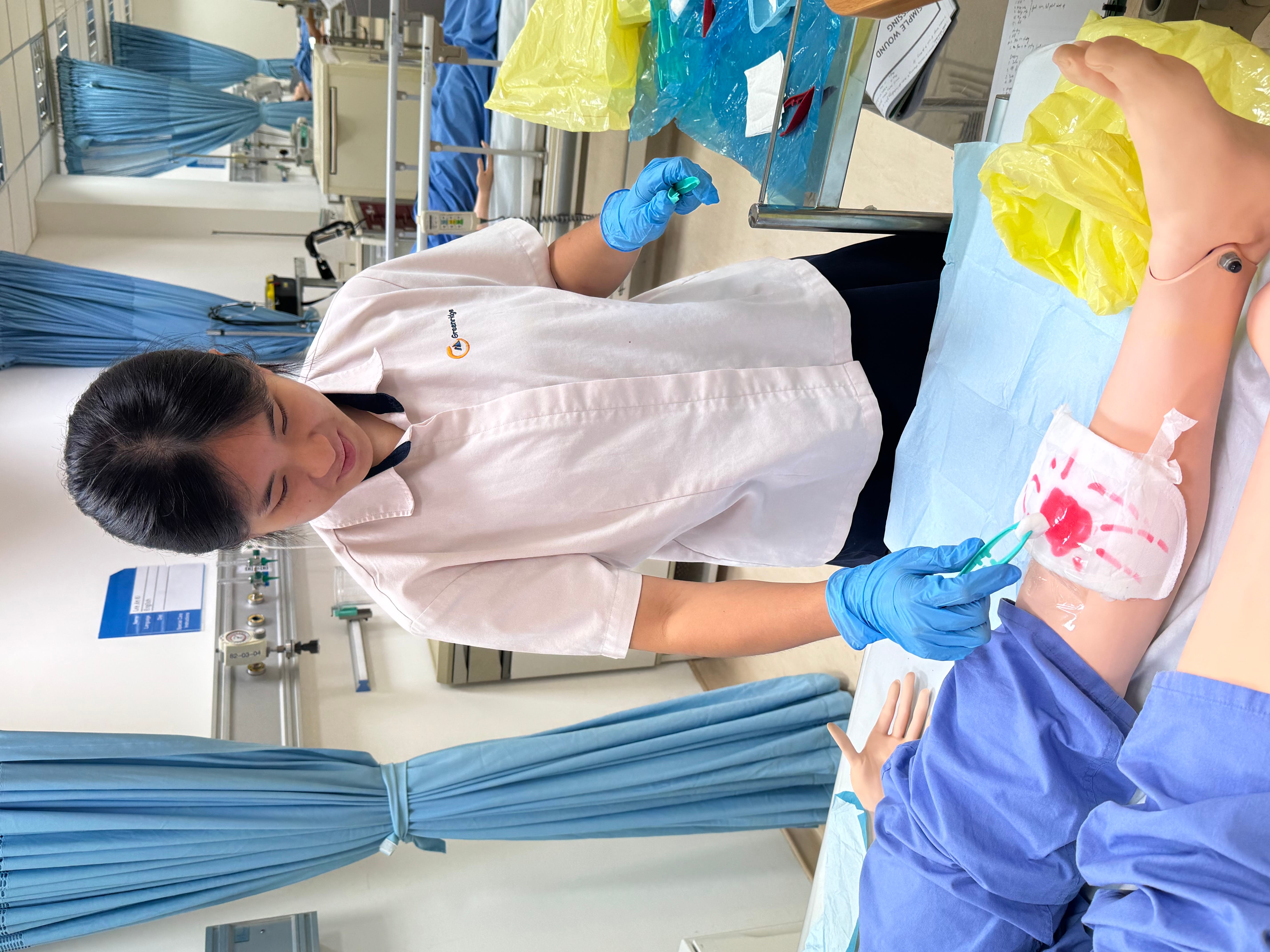 A woman in blue gloves practices wound care on a medical training mannequin leg in a simulation lab.